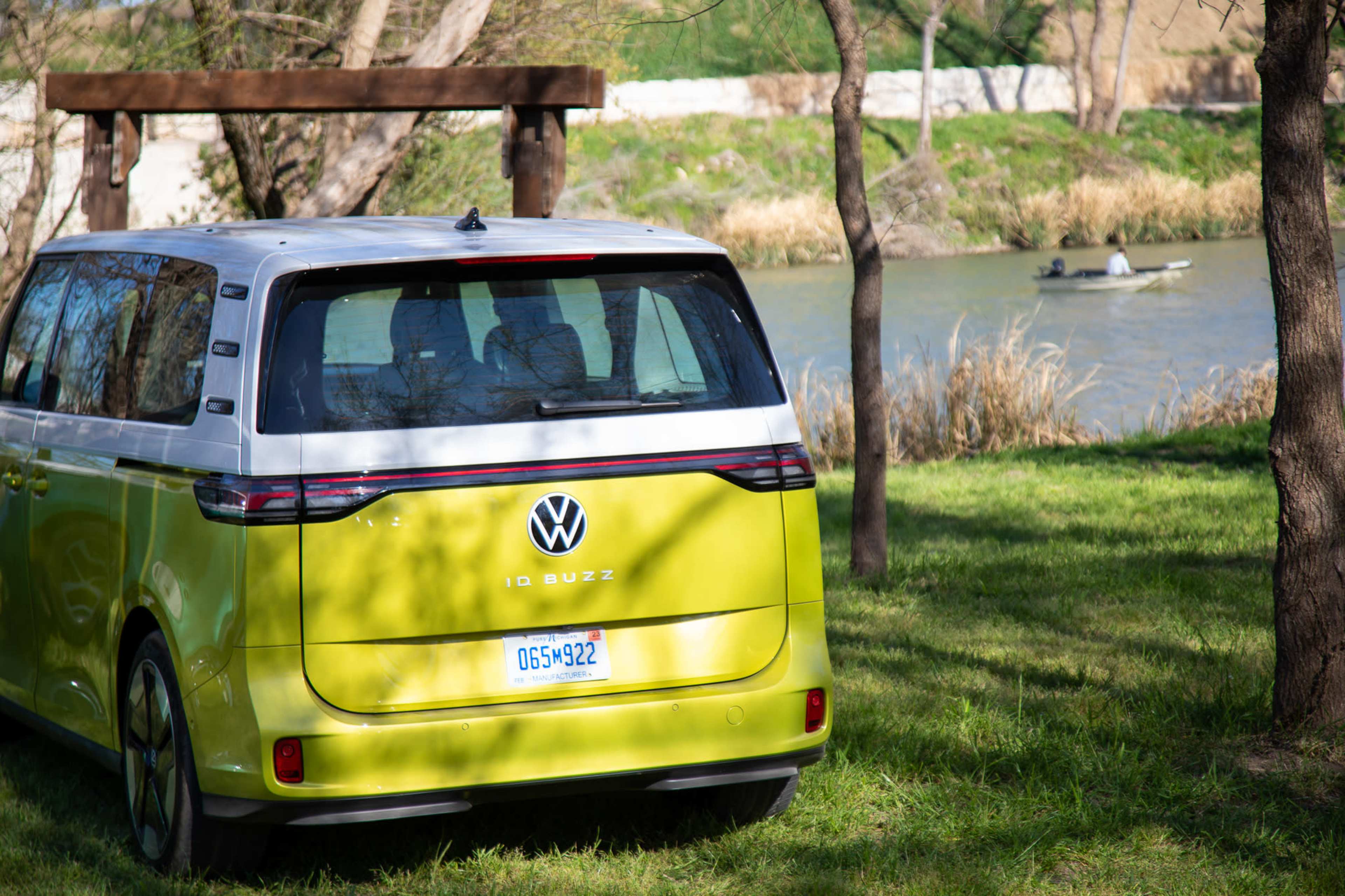 A Volkswagen ID. Buzz van is parked on the grass near a river, with two people in a boat visible in the distance.