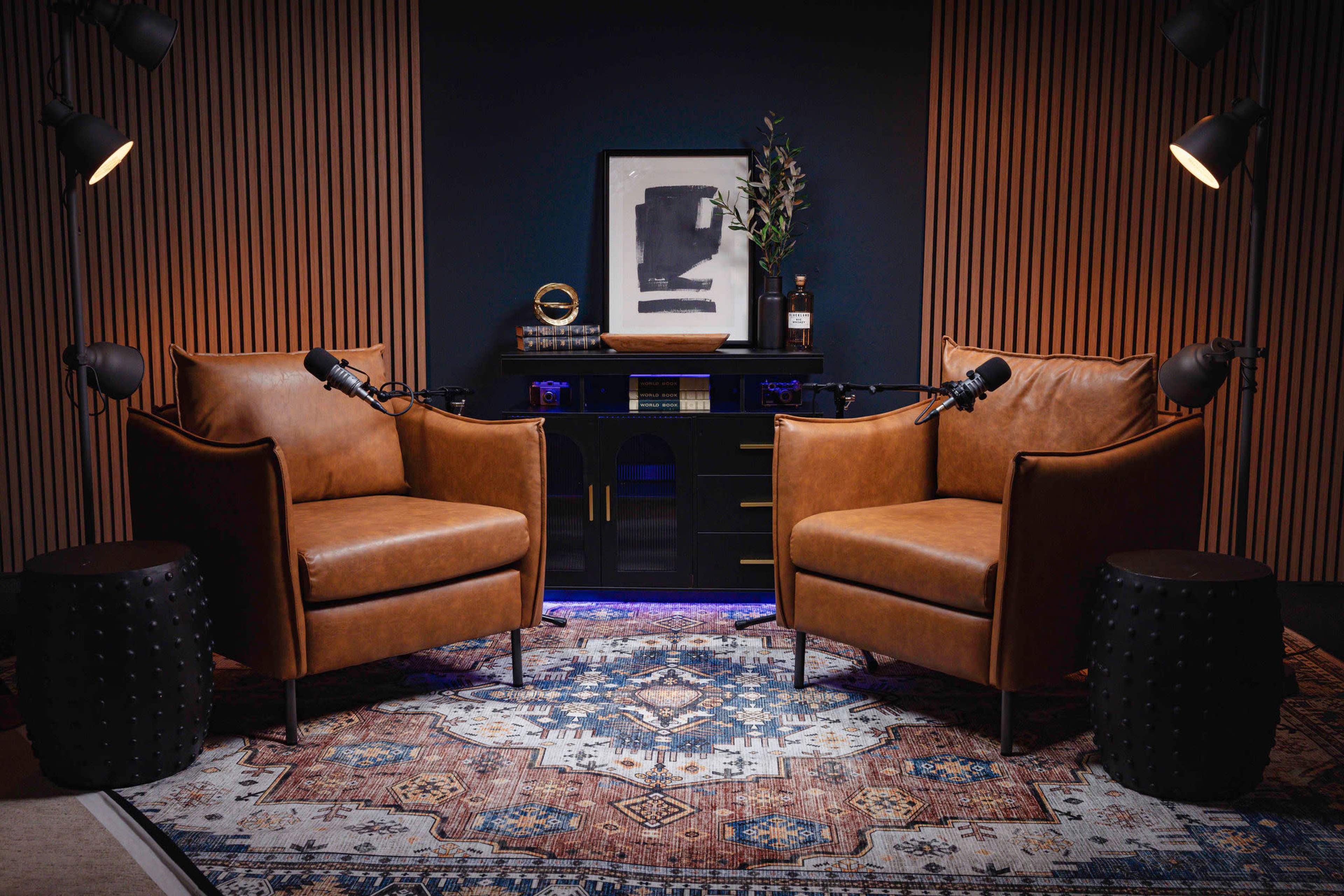 The image shows two brown leather chairs positioned facing each other on a patterned rug, with a dark wood desk and decorative items in the background.