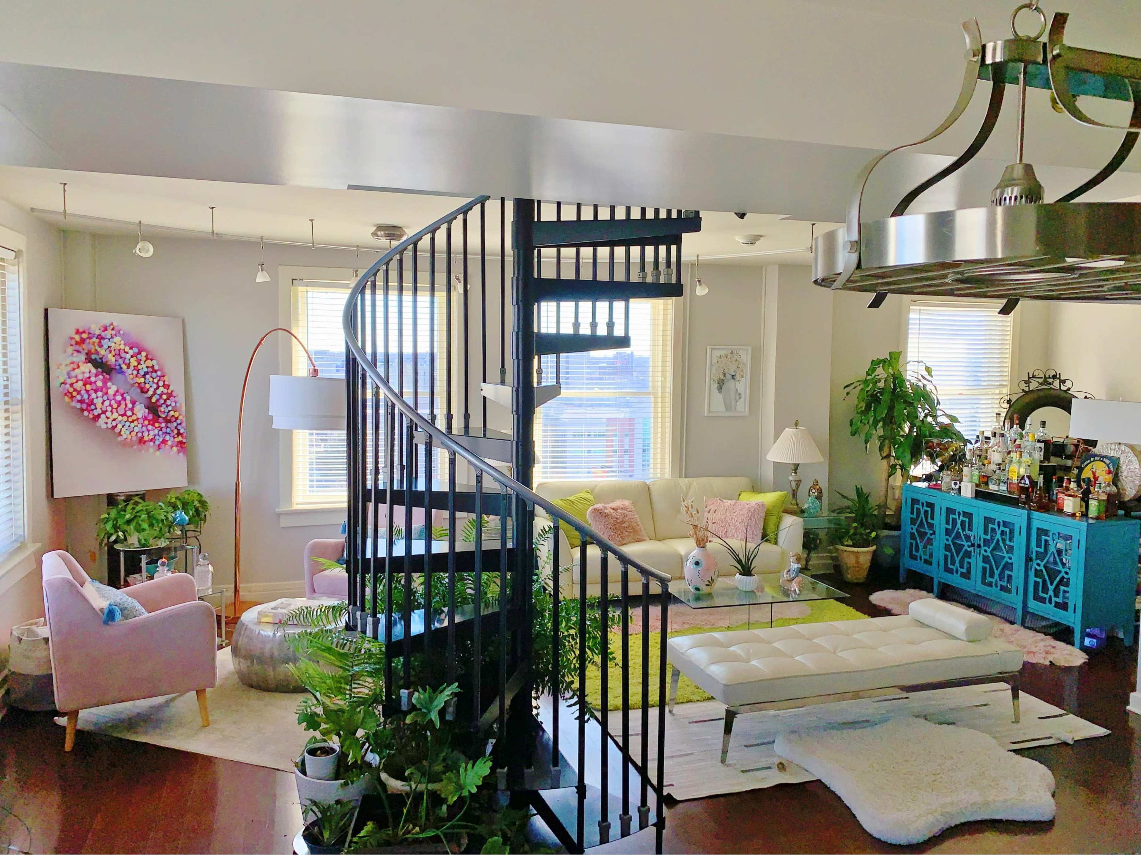 A modern living room with a spiral staircase, featuring colorful furniture, plants, and artistic decor.