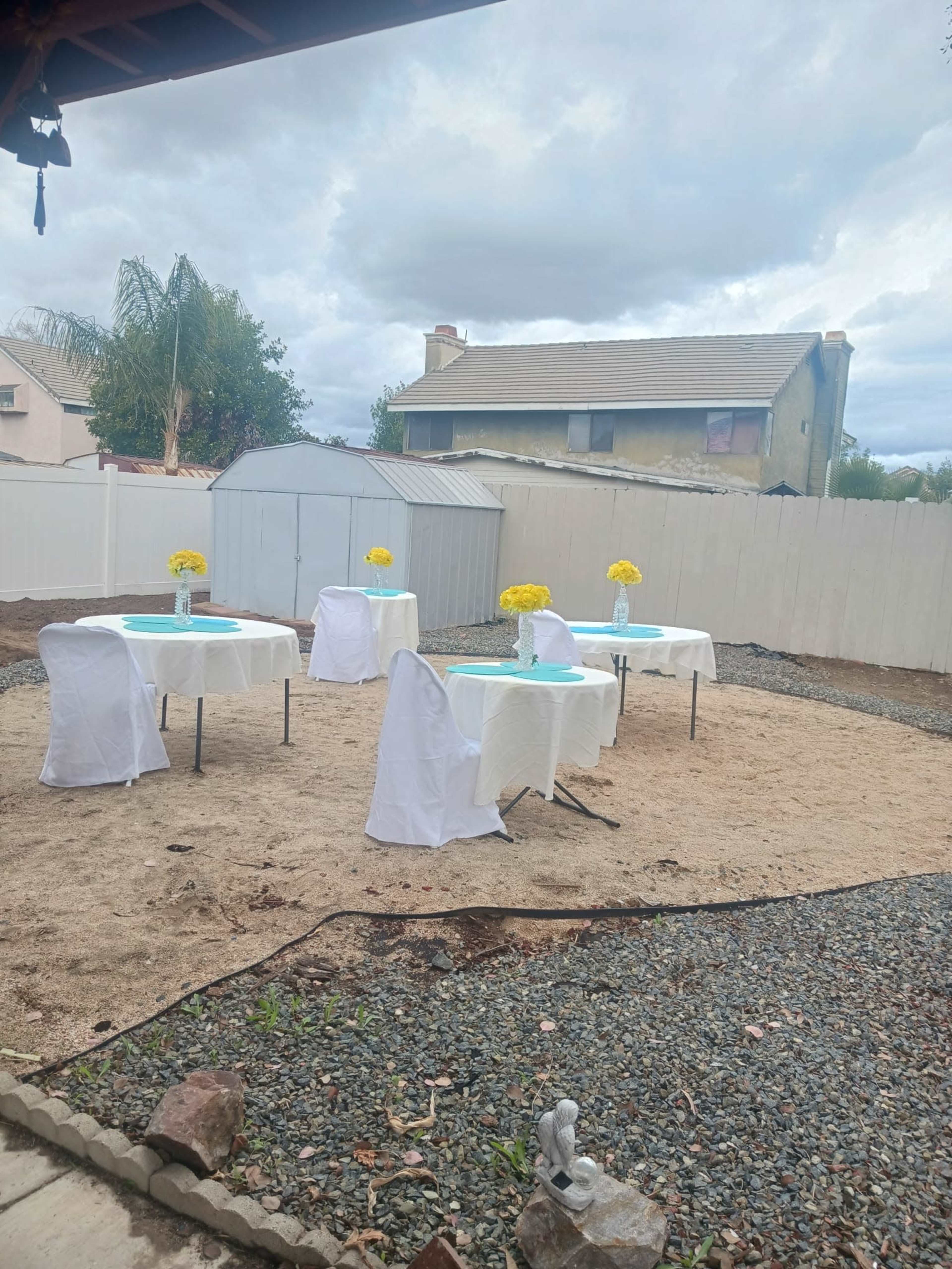 The image shows a backyard setup with four tables covered in white cloths and adorned with yellow flowers, surrounded by a sandy area and a white fence.