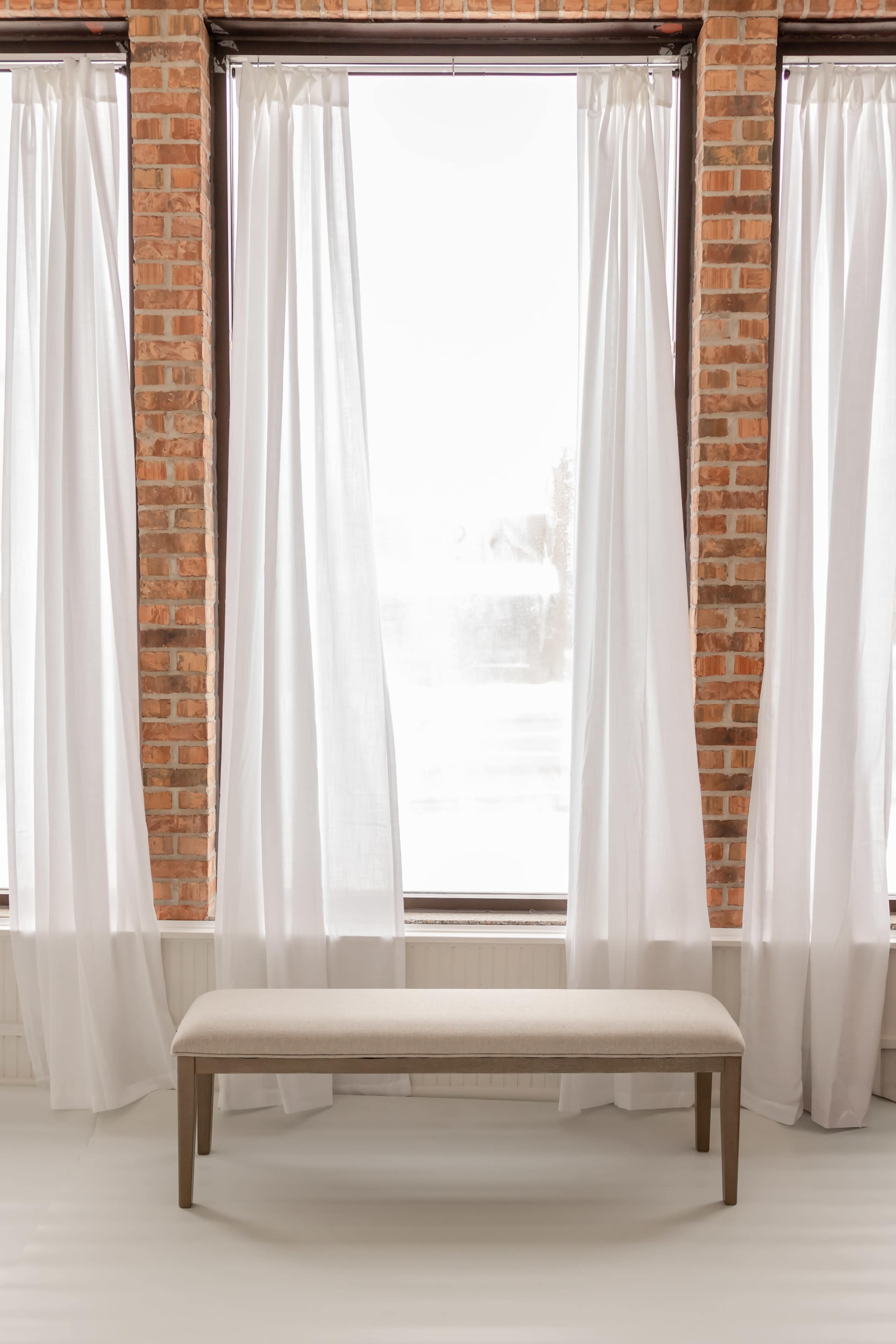 A beige bench is positioned in front of large windows covered with sheer white curtains, framed by exposed brick walls.