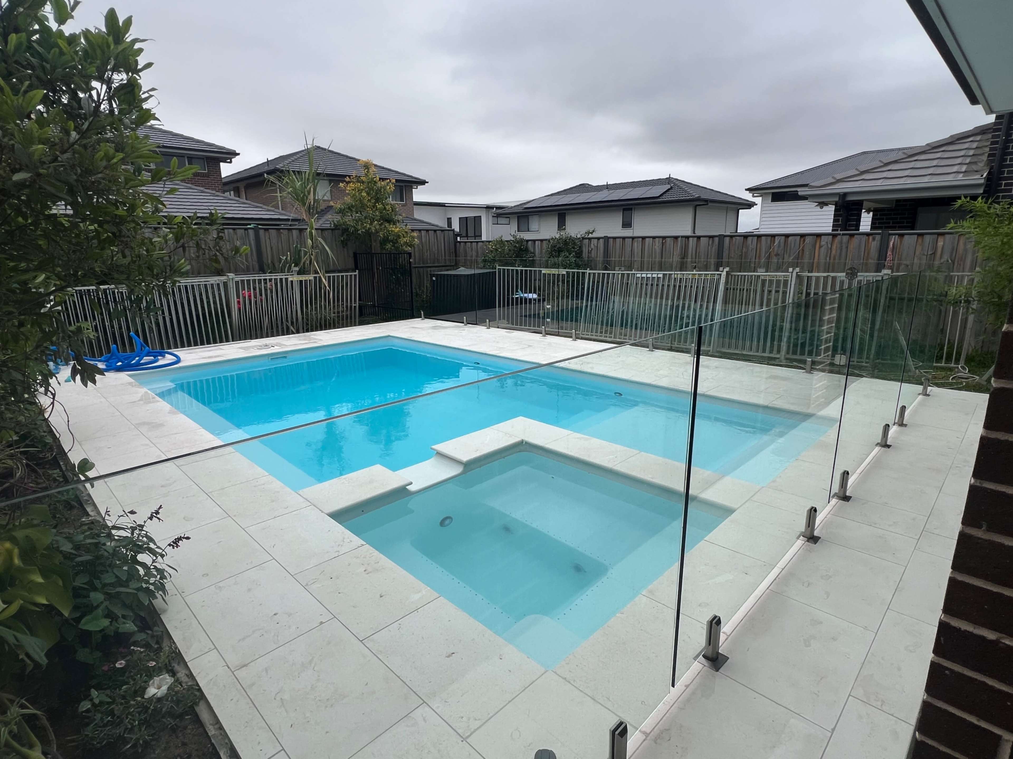 The image shows a modern swimming pool with a hot tub, surrounded by glass fencing and patio pavers in a residential backyard.