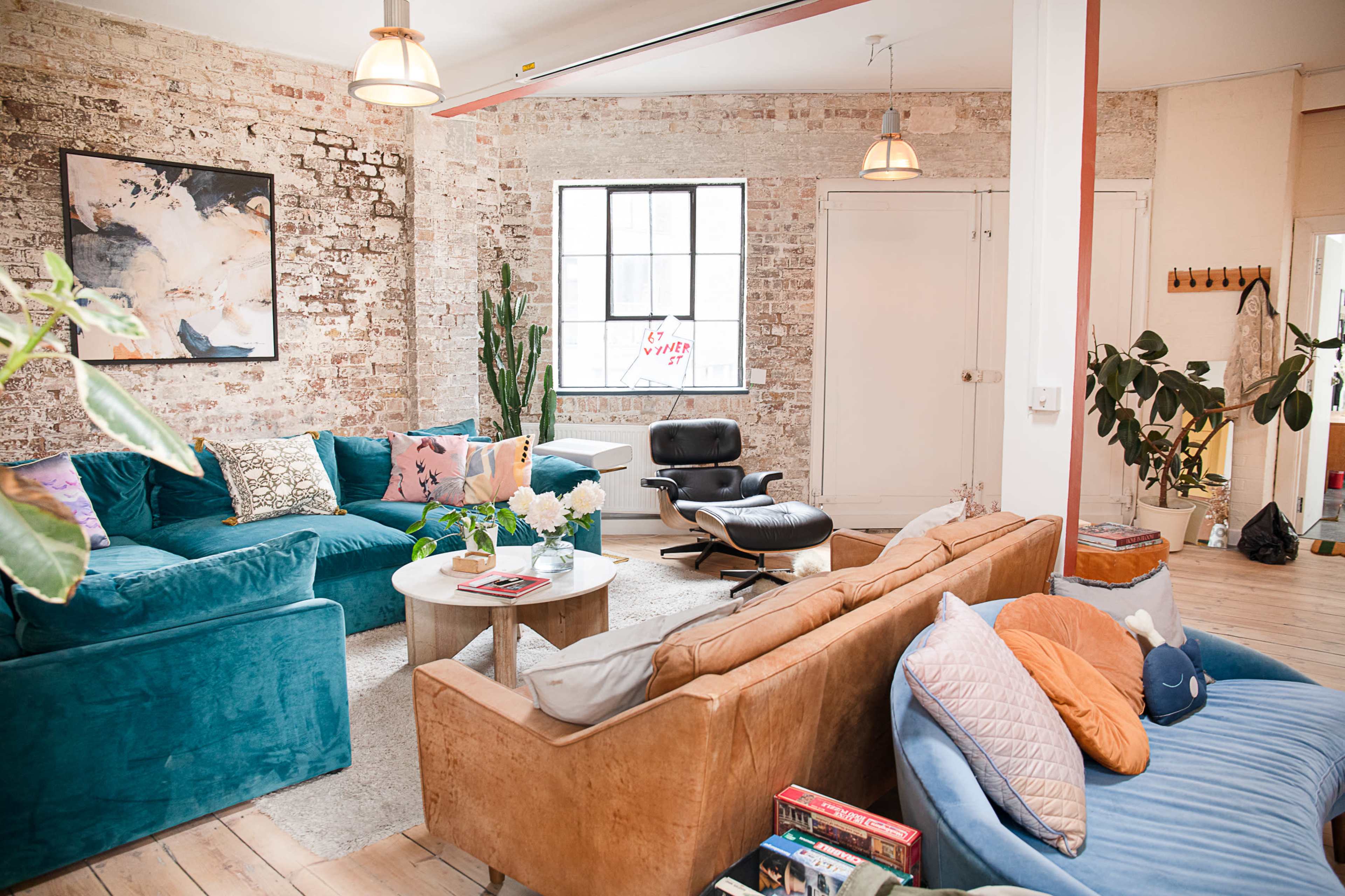 The image shows a cozy living room with exposed brick walls, various seating options including a teal couch and a tan leather sofa, a circular coffee table in the center, and plants throughout the space.