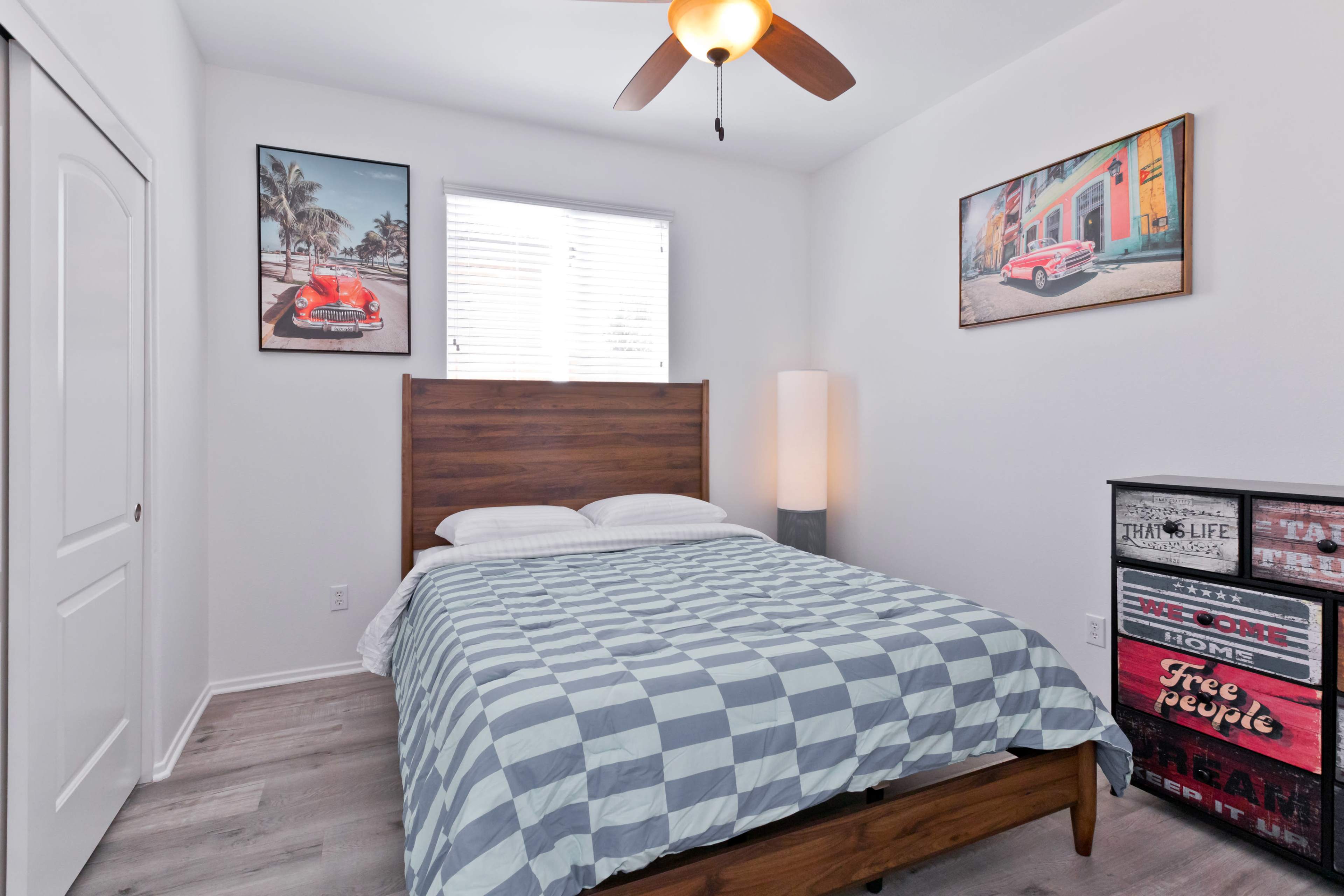 A bedroom features a wooden bed with a checkered blue and white comforter, two framed car posters on the walls, and a lamp beside a storage unit.