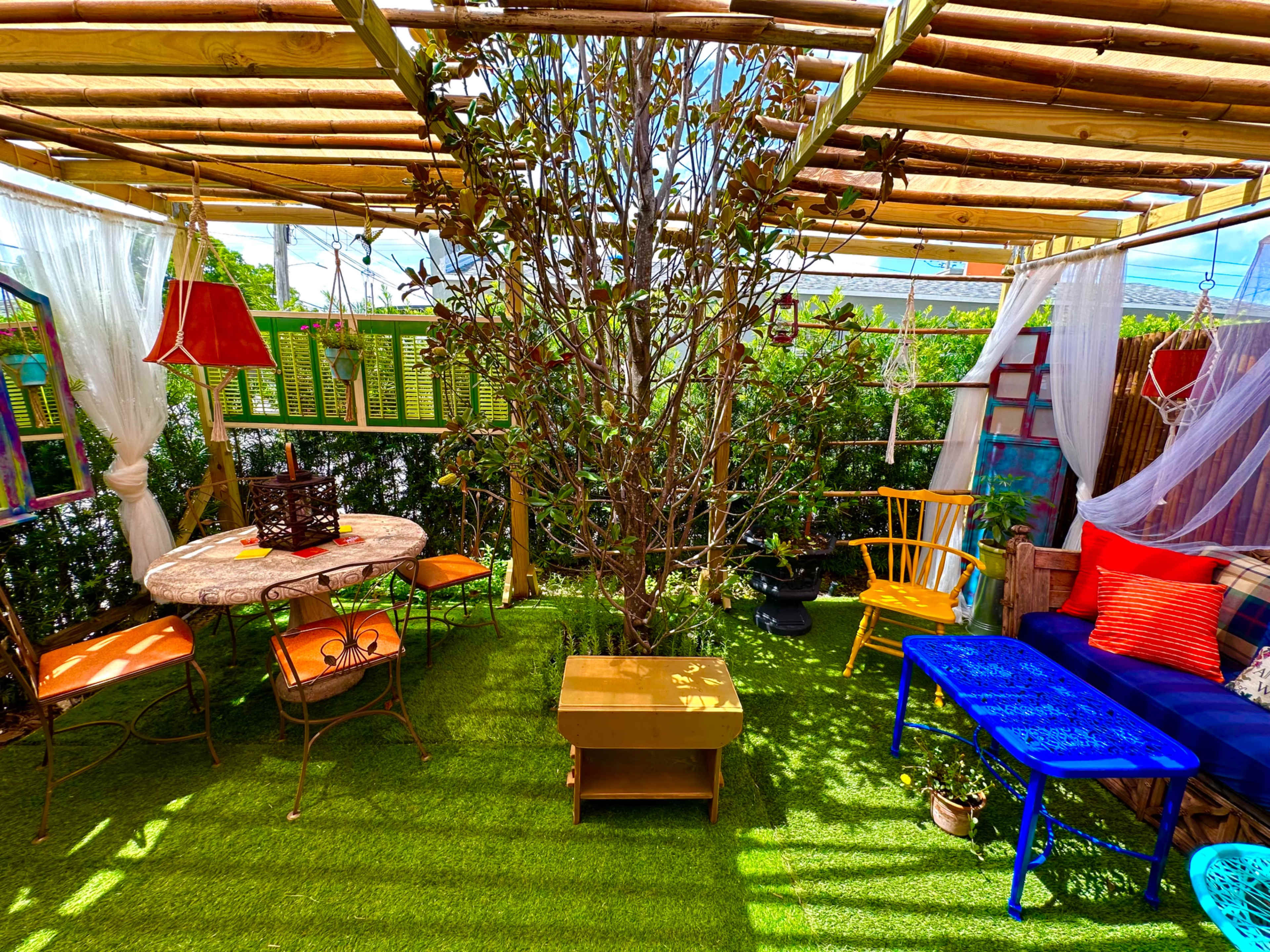 A vibrant outdoor seating area with a table, chairs, a tree, and colorful decor under a bamboo canopy.
