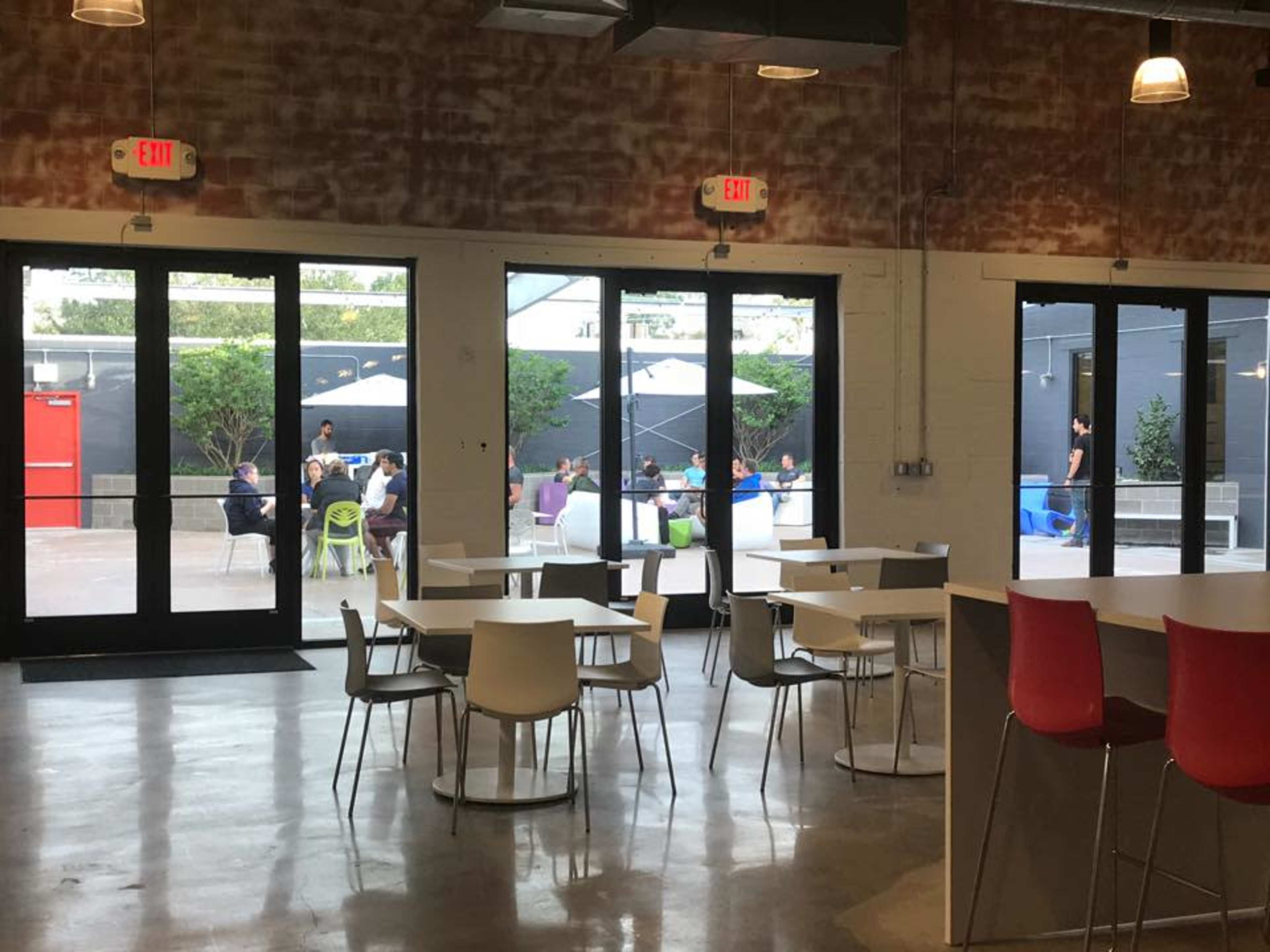 A modern indoor cafe with white and red furniture, large glass doors opening to an outdoor seating area where people are gathered under umbrellas.