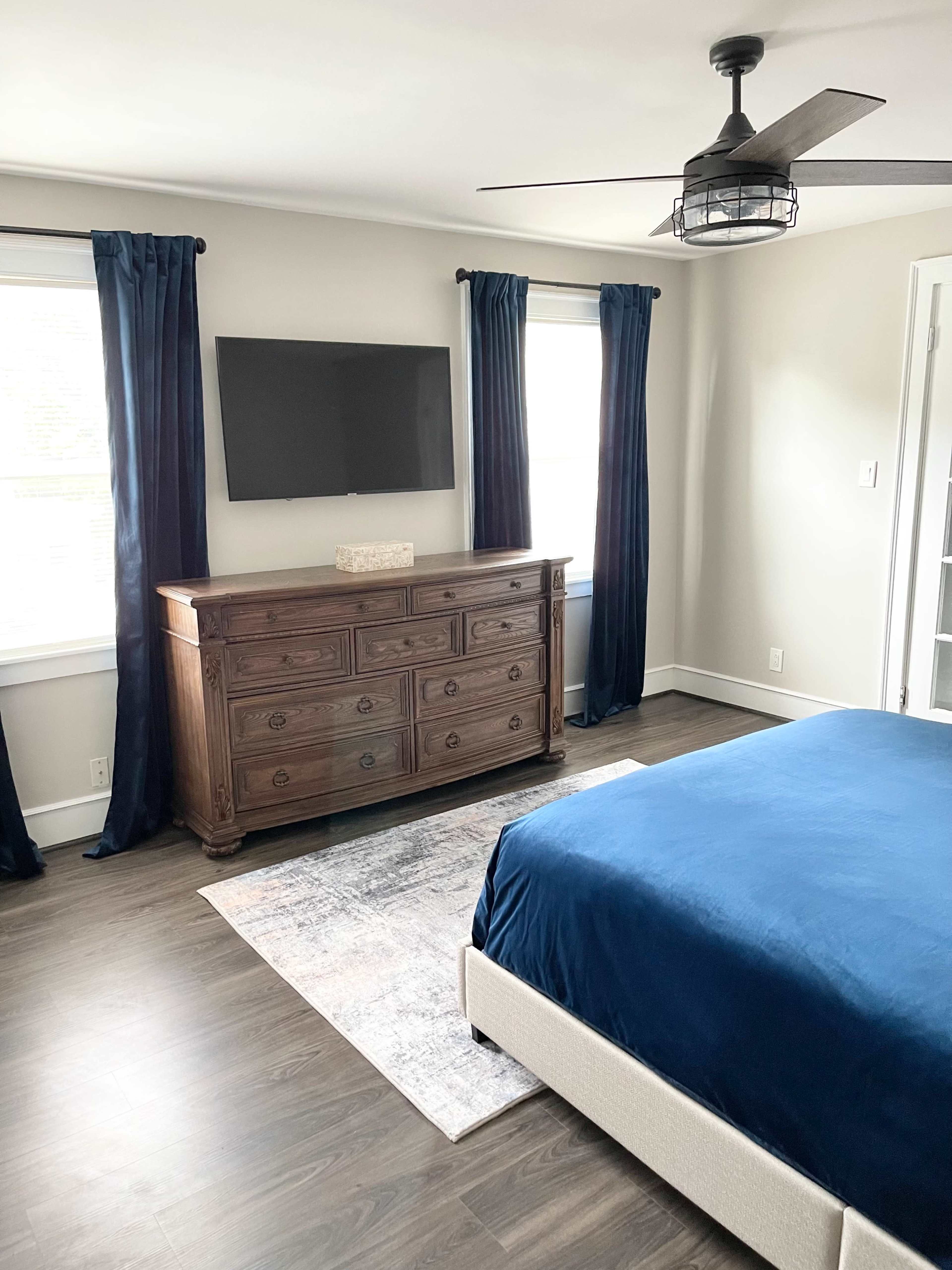 A modern bedroom with a blue bedspread, a wooden dresser, a wall-mounted television, and large windows covered with dark curtains.
