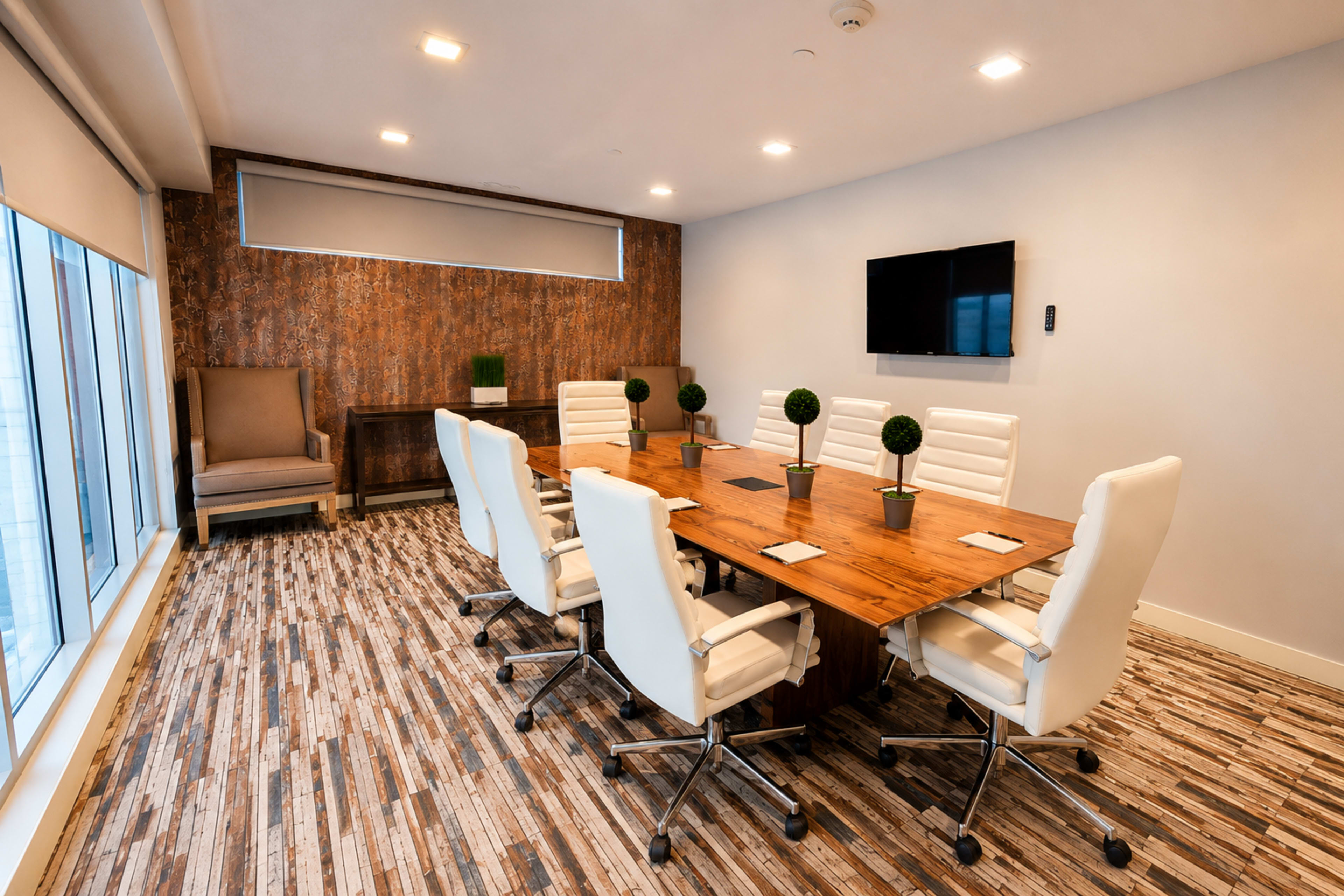 The image shows a modern conference room featuring a large wooden table surrounded by white ergonomic chairs, with a wall-mounted TV and decorative plants on the table.