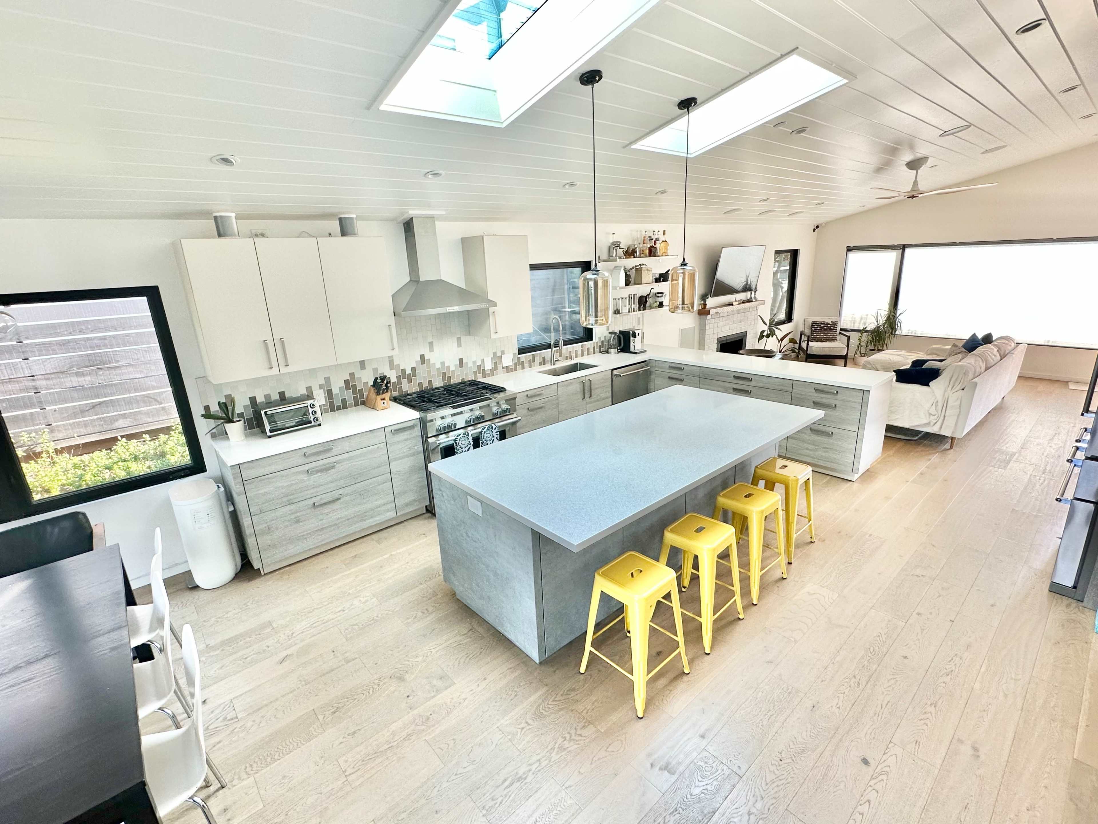 A modern kitchen features a large island with yellow stools, stainless steel appliances, and a spacious layout defined by minimalistic cabinetry and an abundance of natural light.