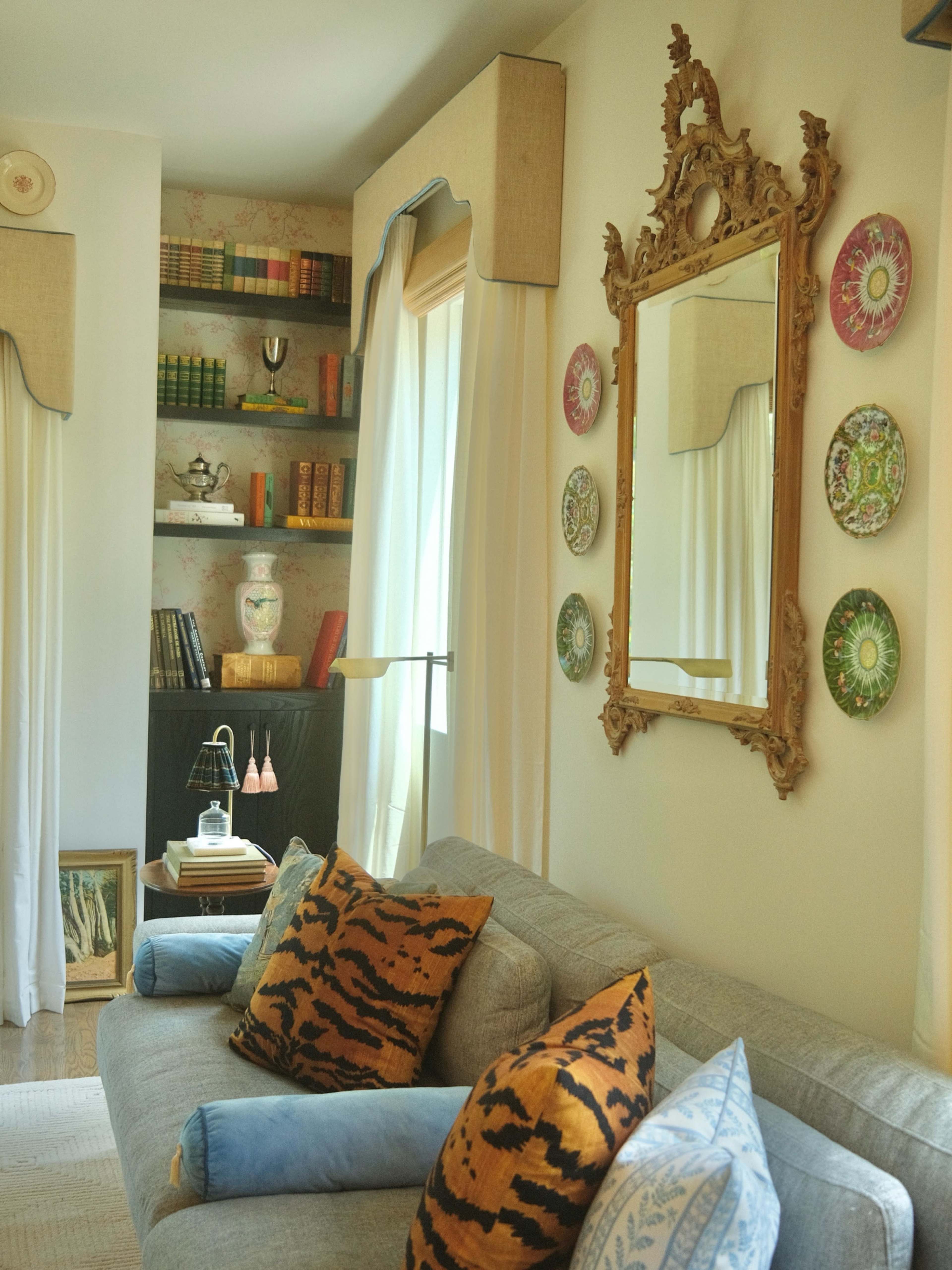 The image shows a cozy living room with a gray sofa adorned with patterned pillows, a decorative mirror on the wall, and shelves lined with books and ornaments.