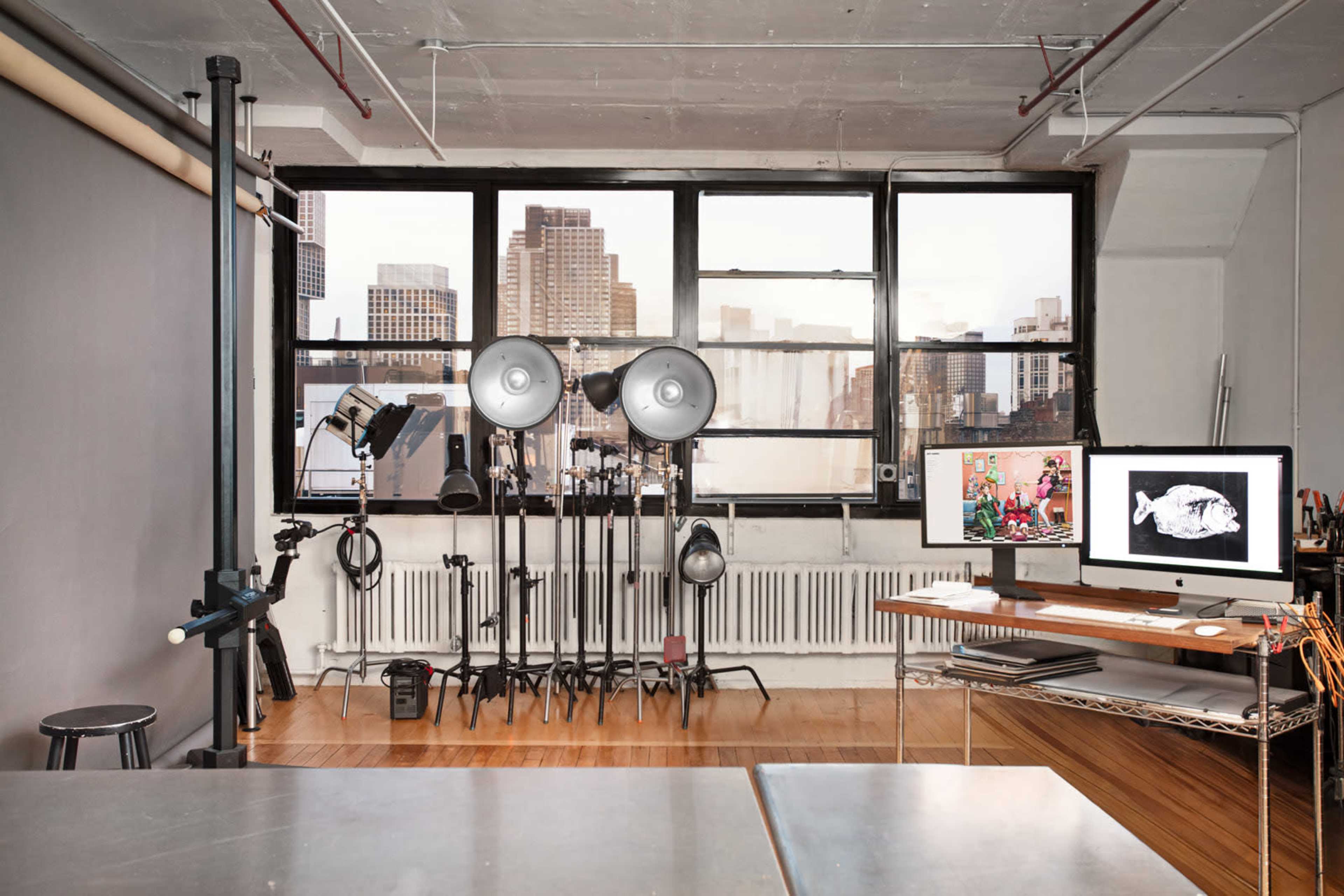 The image shows a photography studio with large windows overlooking a cityscape, equipped with lighting equipment and a desk featuring a computer monitor.