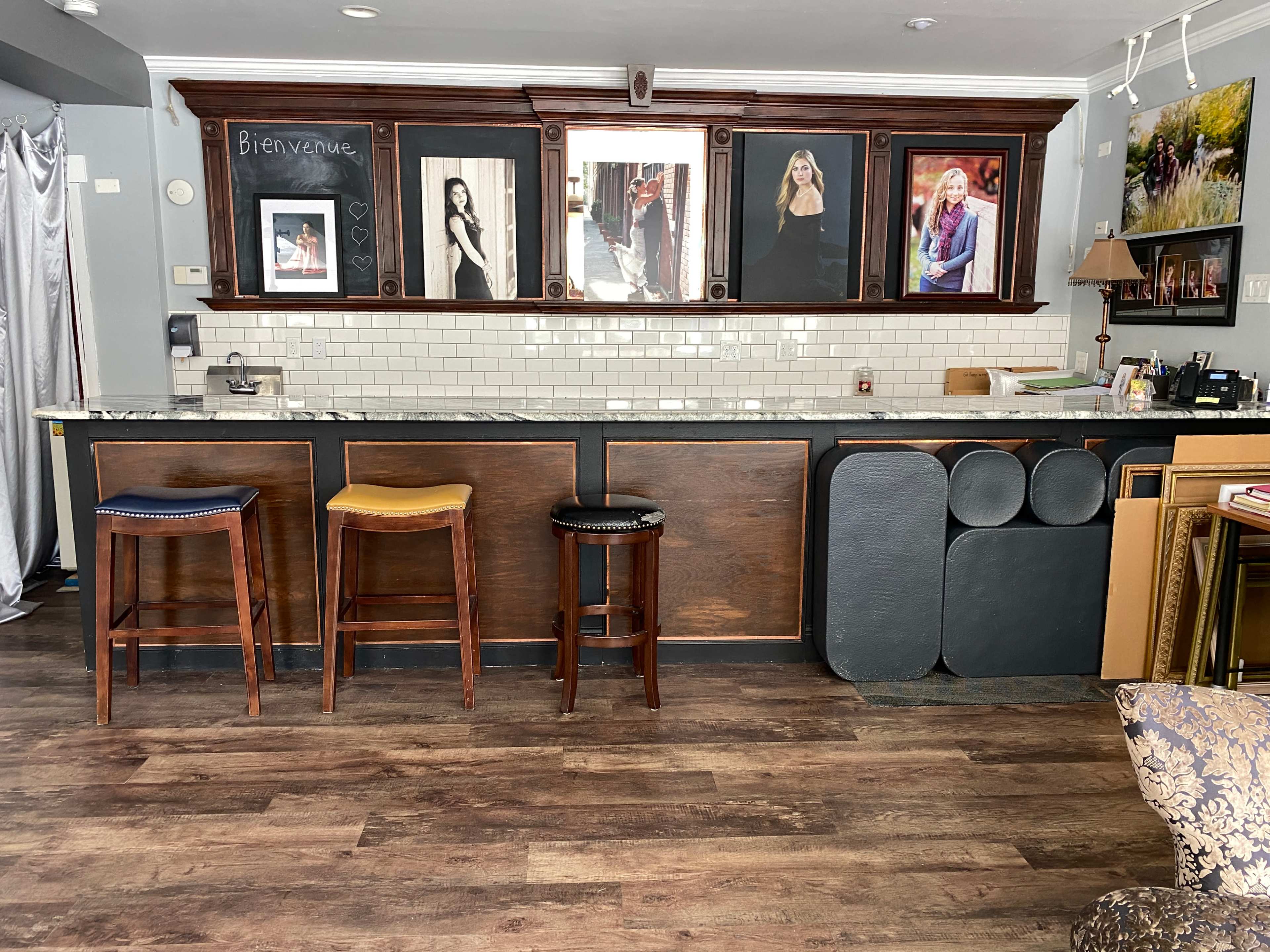 The image shows a bar area with three colorful stools in front of a wooden counter, adorned with framed photographs on the wall behind it.