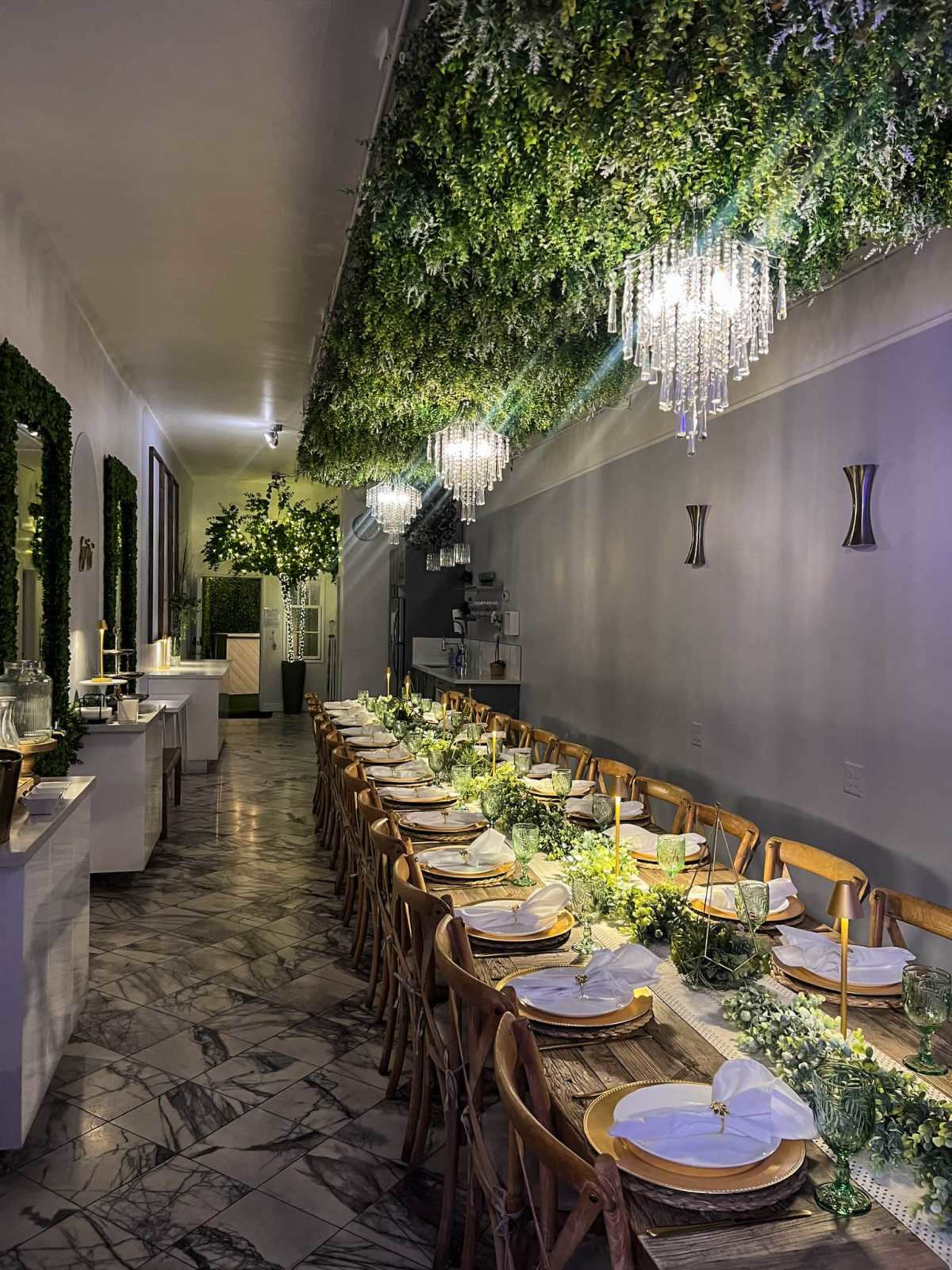 A long dining table is set in an elegantly decorated room with greenery overhead and crystal chandeliers.