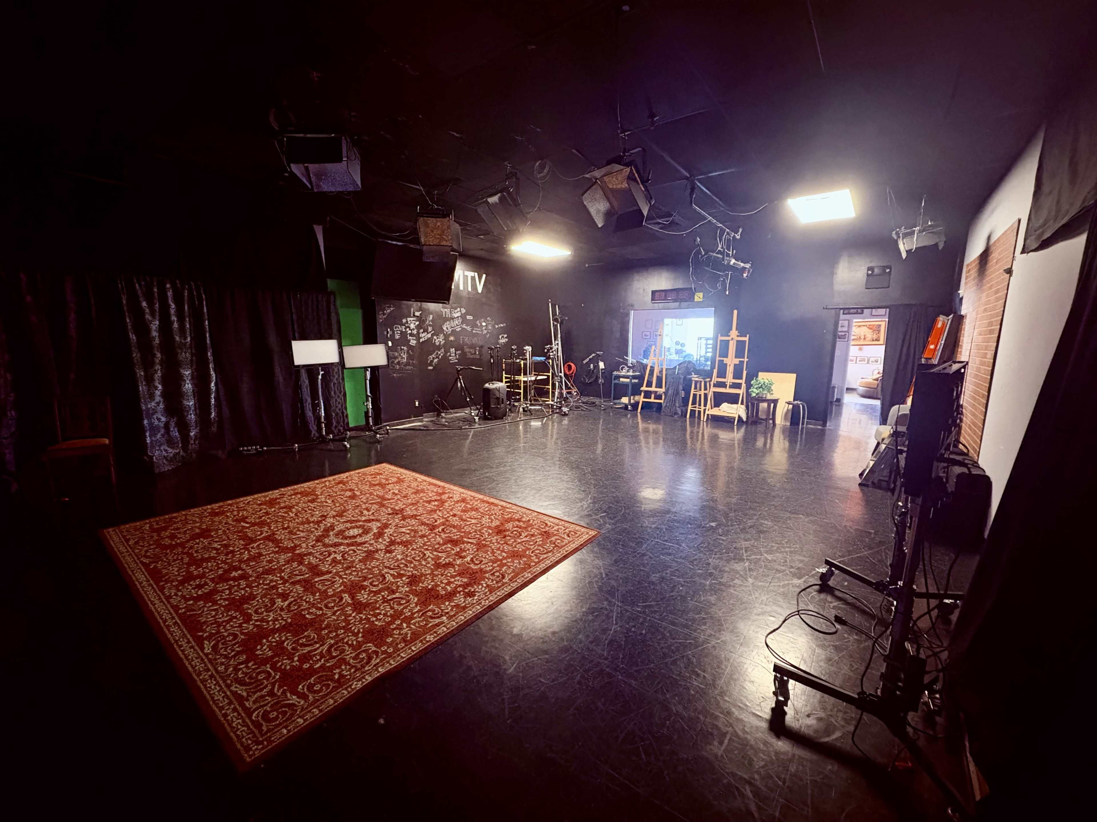 The image shows a dimly lit studio space with a patterned rug, scattered lighting equipment, and various props arranged around a center stage area.