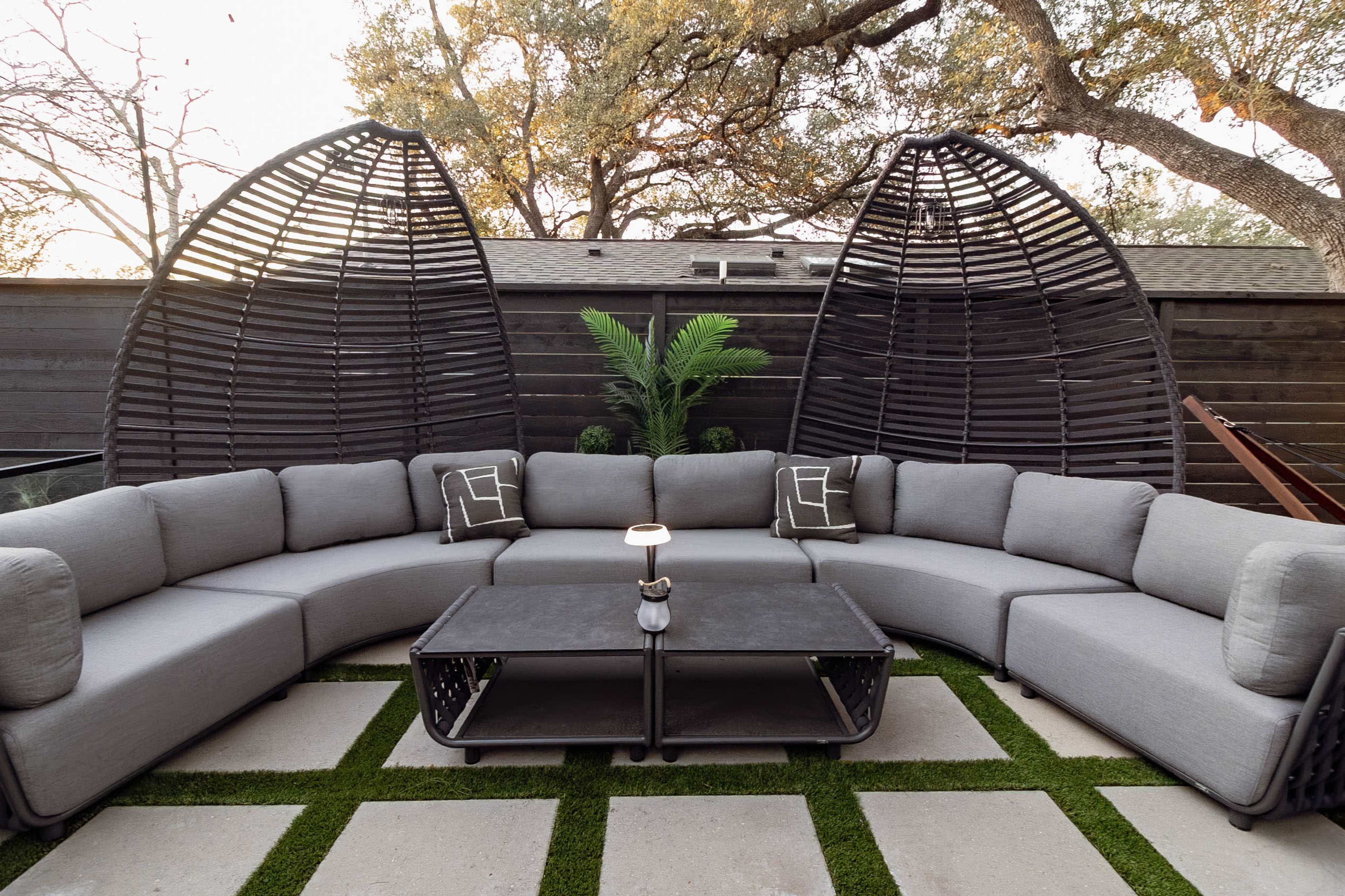 The image shows a modern outdoor seating area featuring a curved gray sectional sofa and a black coffee table surrounded by greenery and decorative structures.