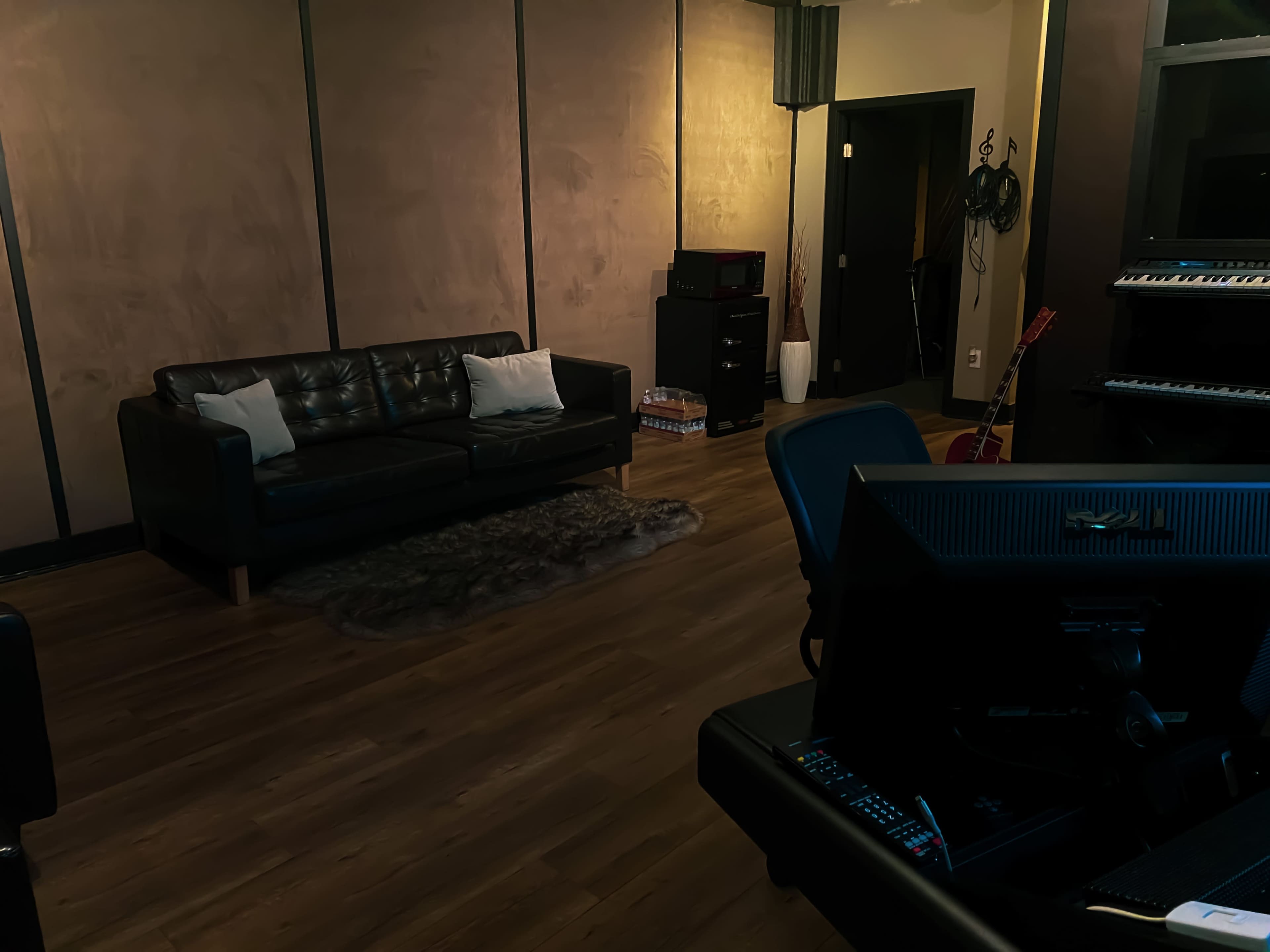 A dimly lit music studio featuring a black sofa, a rug, a computer desk, and various musical instruments in the background.