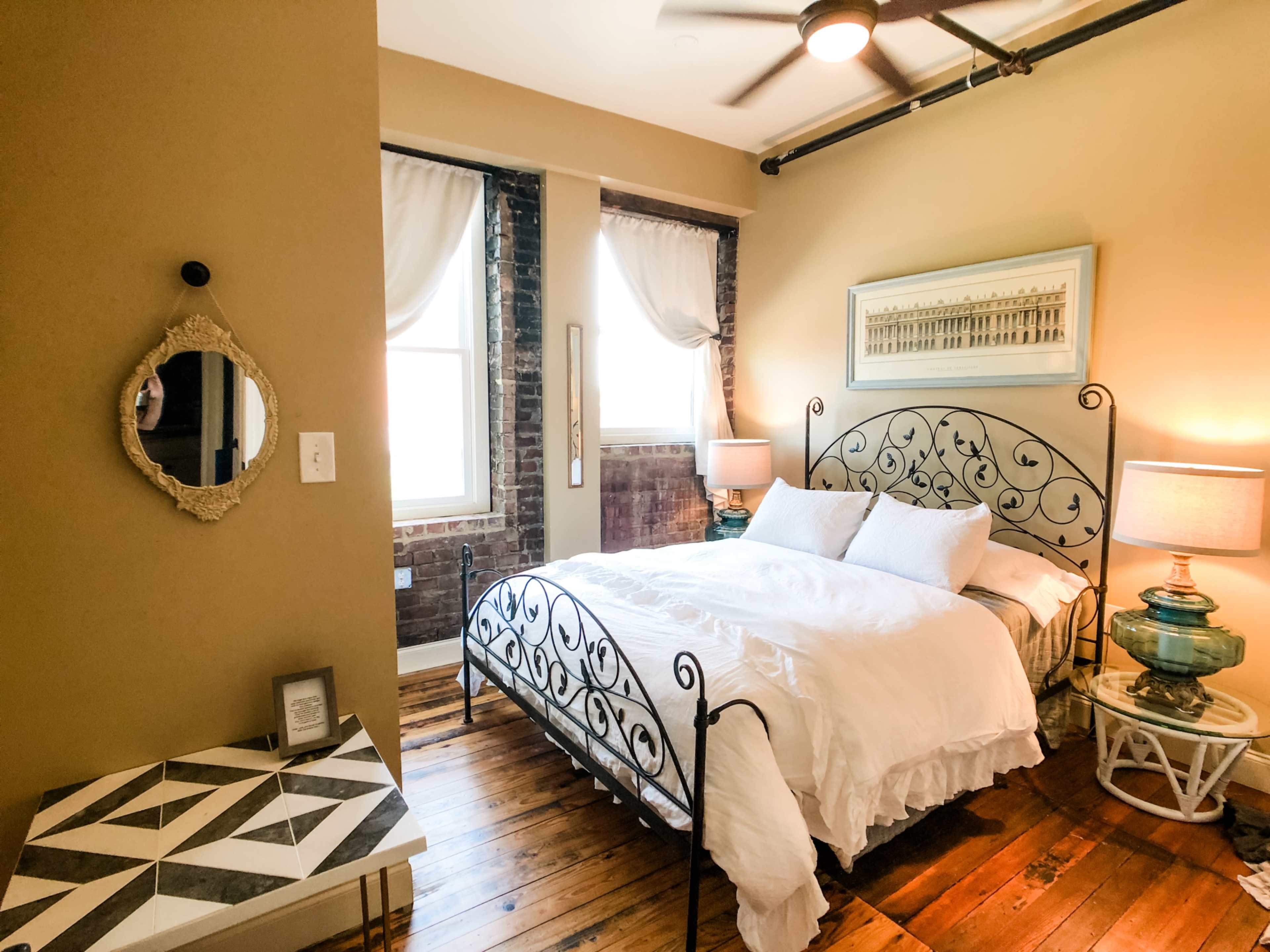 A cozy bedroom with a wrought iron bed frame, two bedside lamps, a decorative mirror, and exposed brick walls.