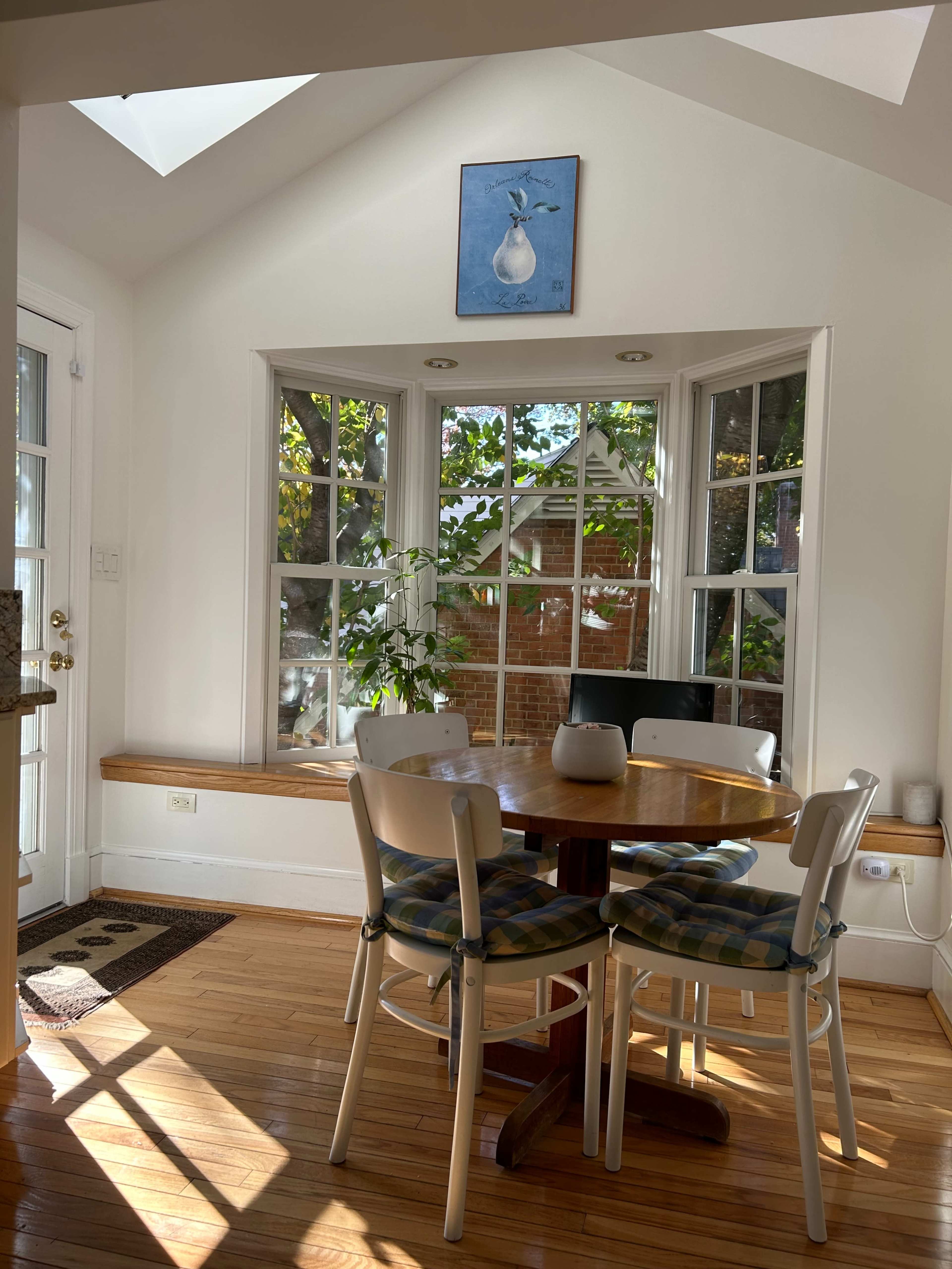 A sunlit dining area features a round wooden table surrounded by four chairs, with a painting of a pear hanging on the wall above.