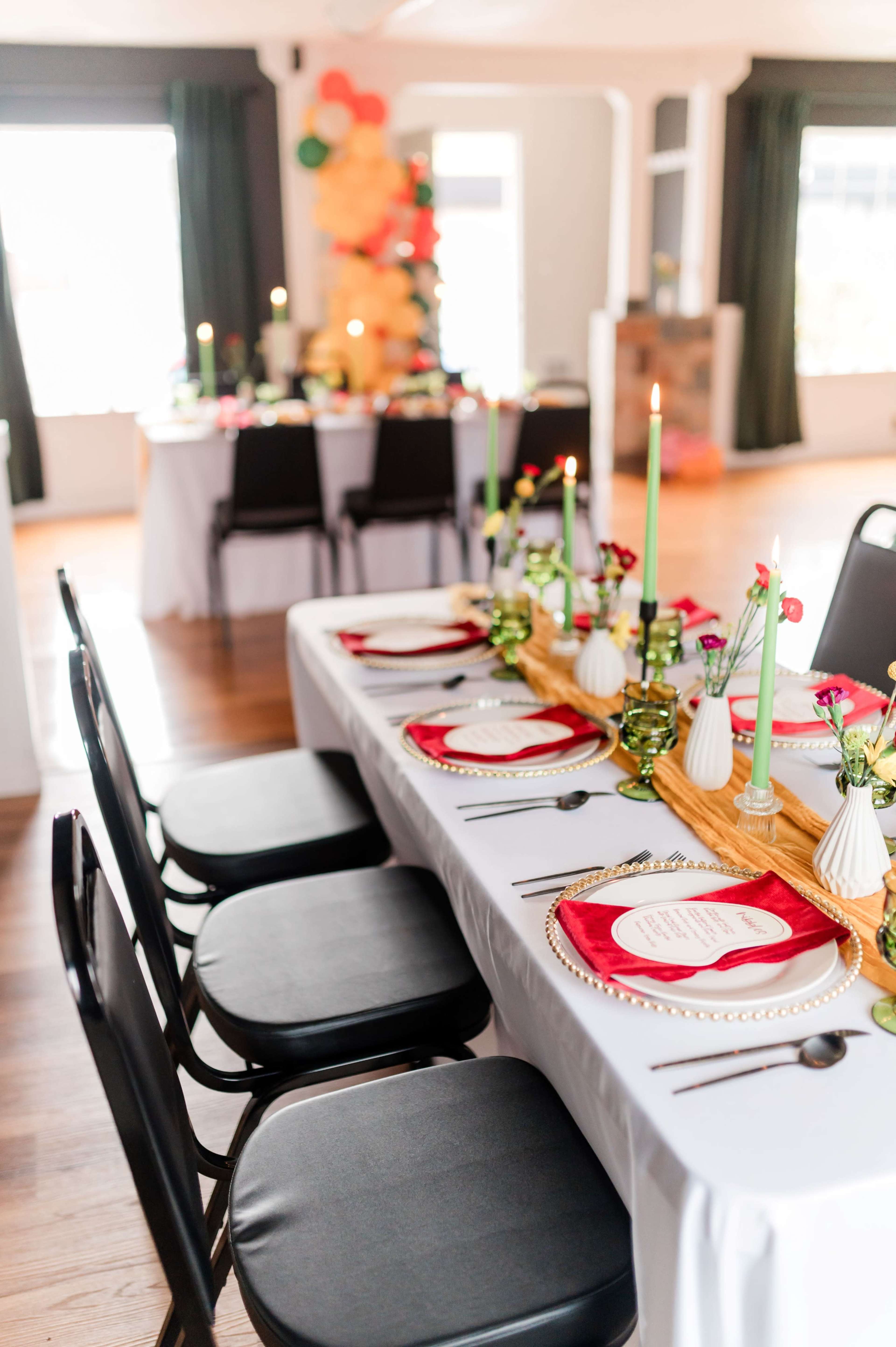 A long dining table is set with red plates, green candles, and decorative centerpieces, while a second table with a festive arrangement is visible in the background.