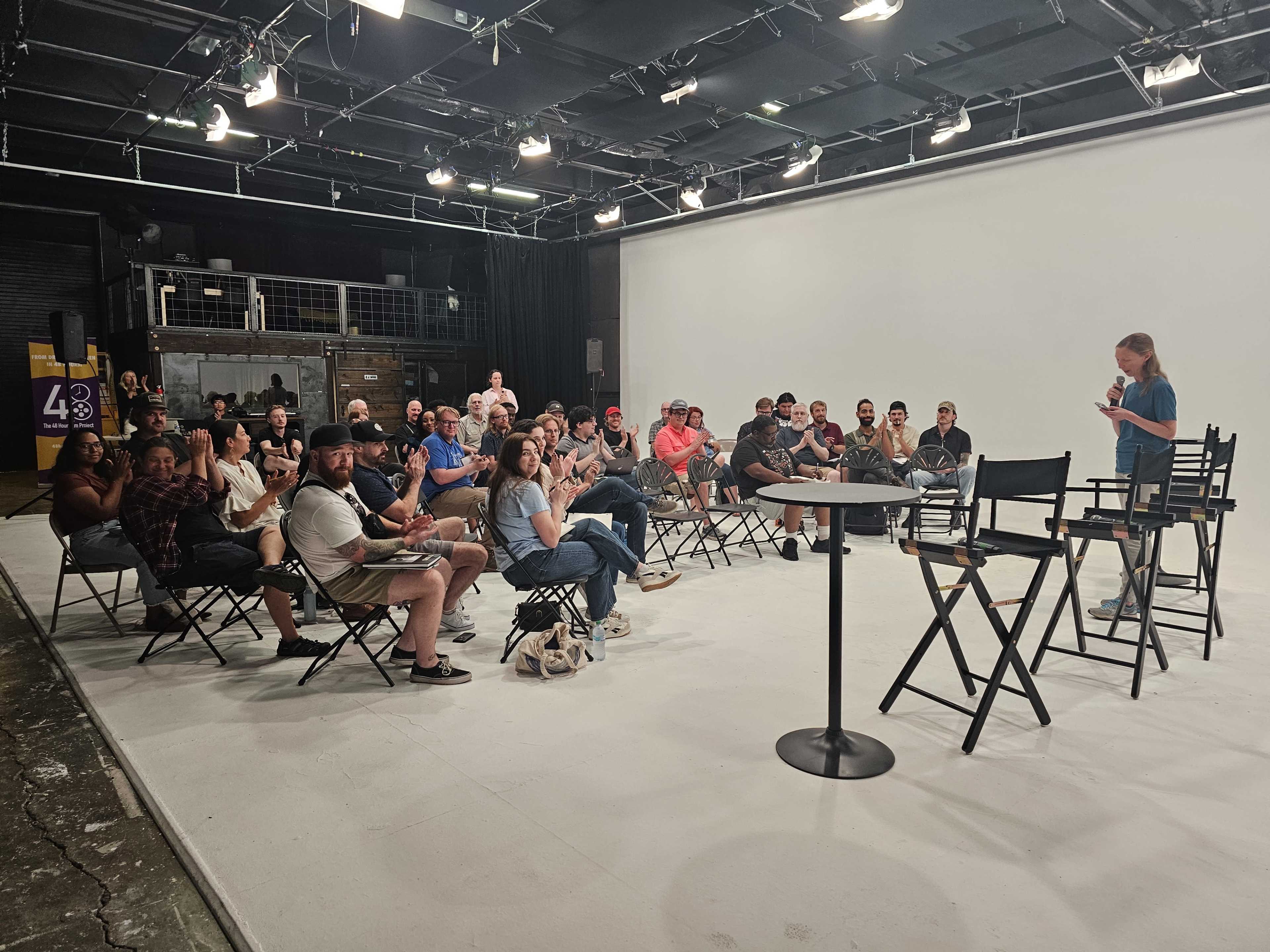 A diverse group of people sits in folding chairs facing a speaker on a stage in a well-lit studio.
