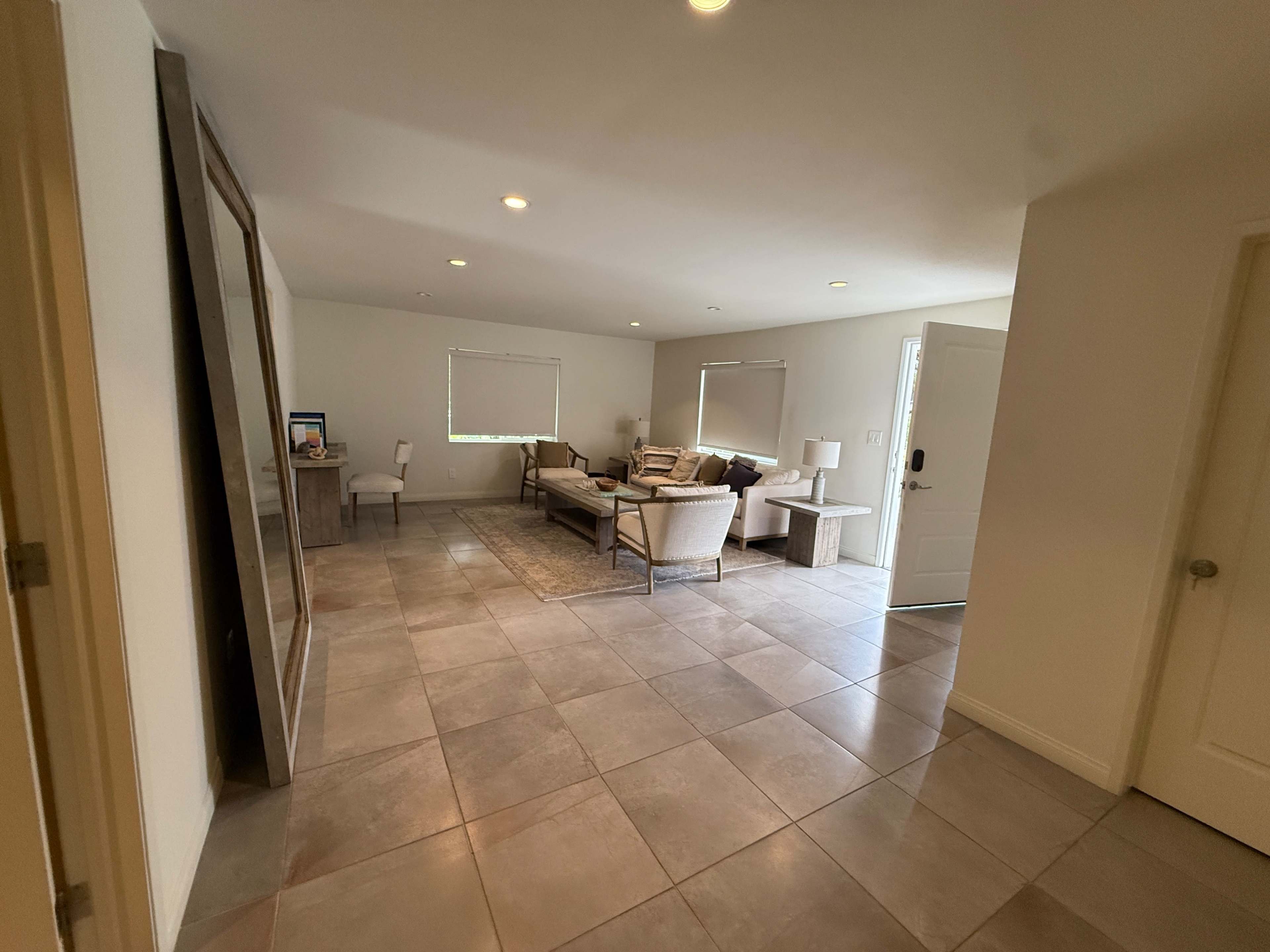 The image shows a spacious living room with tile flooring, featuring a sofa set, a small table, and two chairs near a window, with a door leading outside.