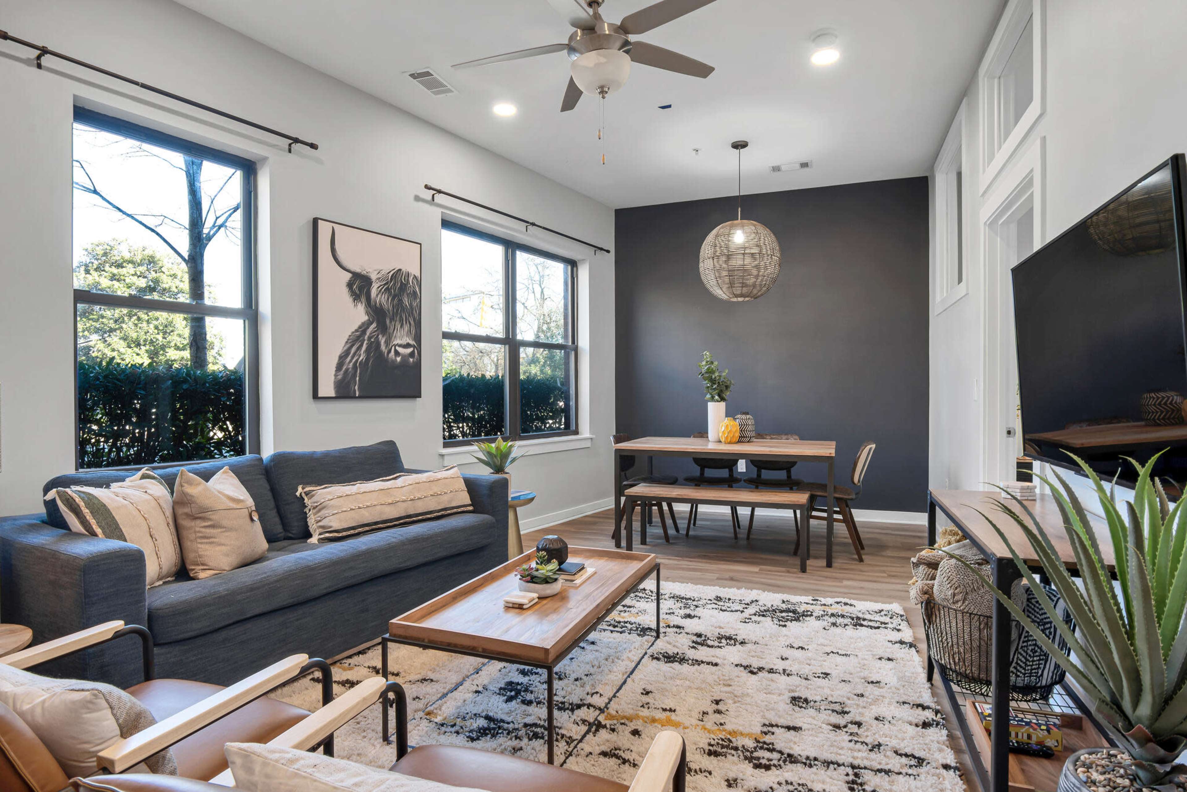 The image shows a modern living room with a blue sofa, a wooden dining table, and a large wall art piece featuring a cow, set against a dark feature wall.
