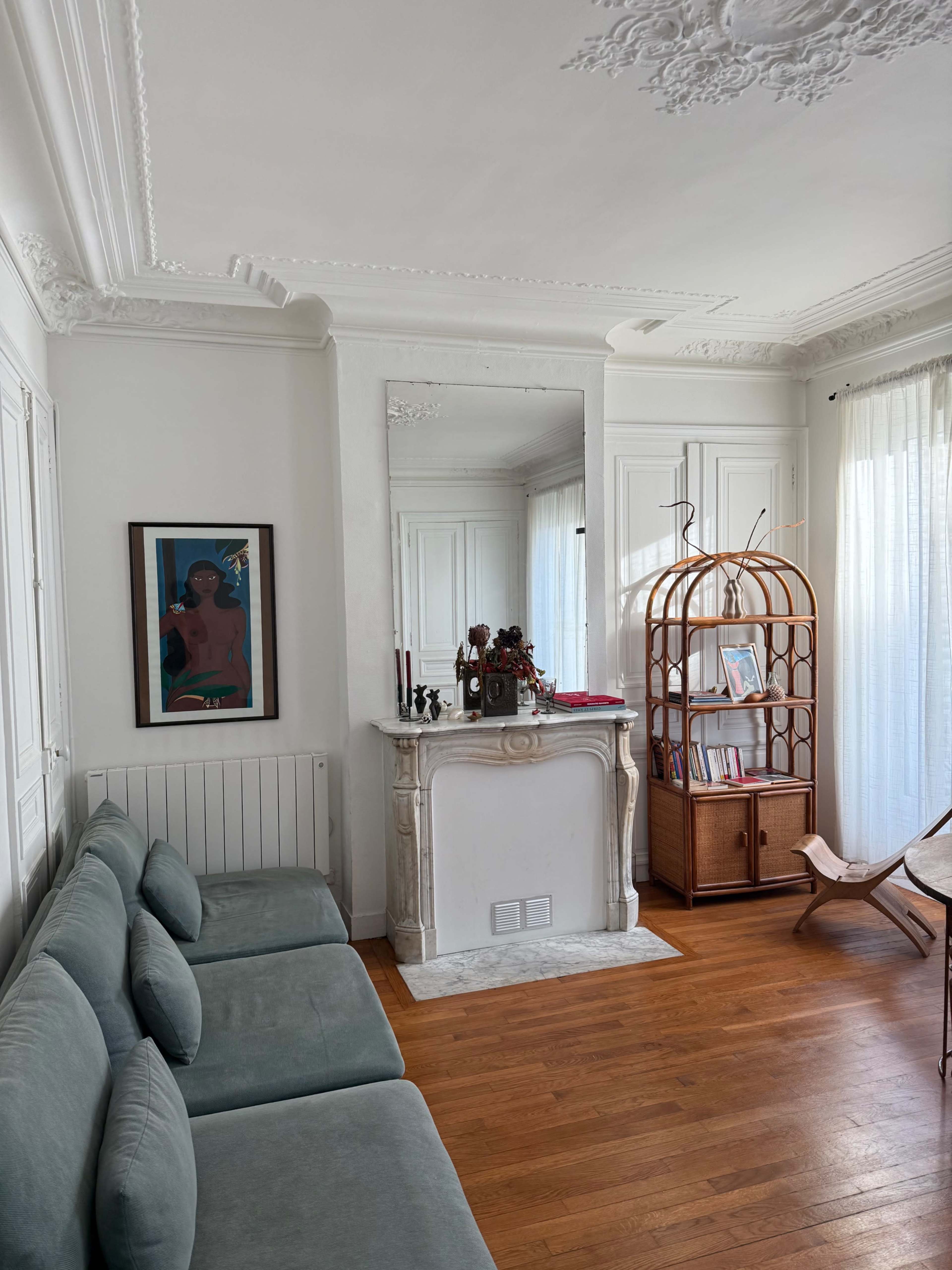 The image shows a bright living room featuring a light-colored couch, a decorative fireplace with a mirror above it, a wicker bookshelf, and a framed artwork on the wall.