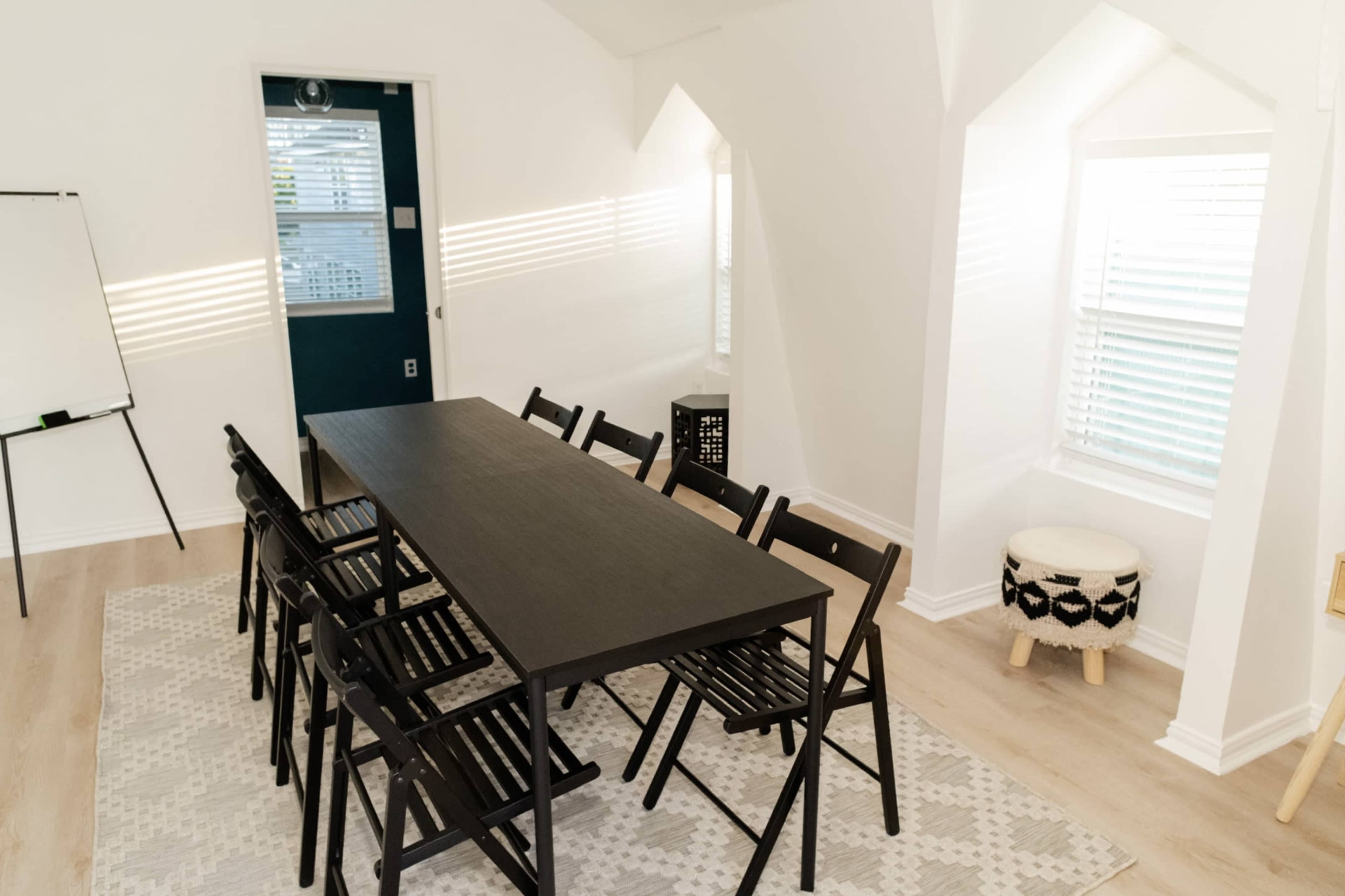 A long black table with eight chairs is set in a bright room with large windows and minimalist decor.