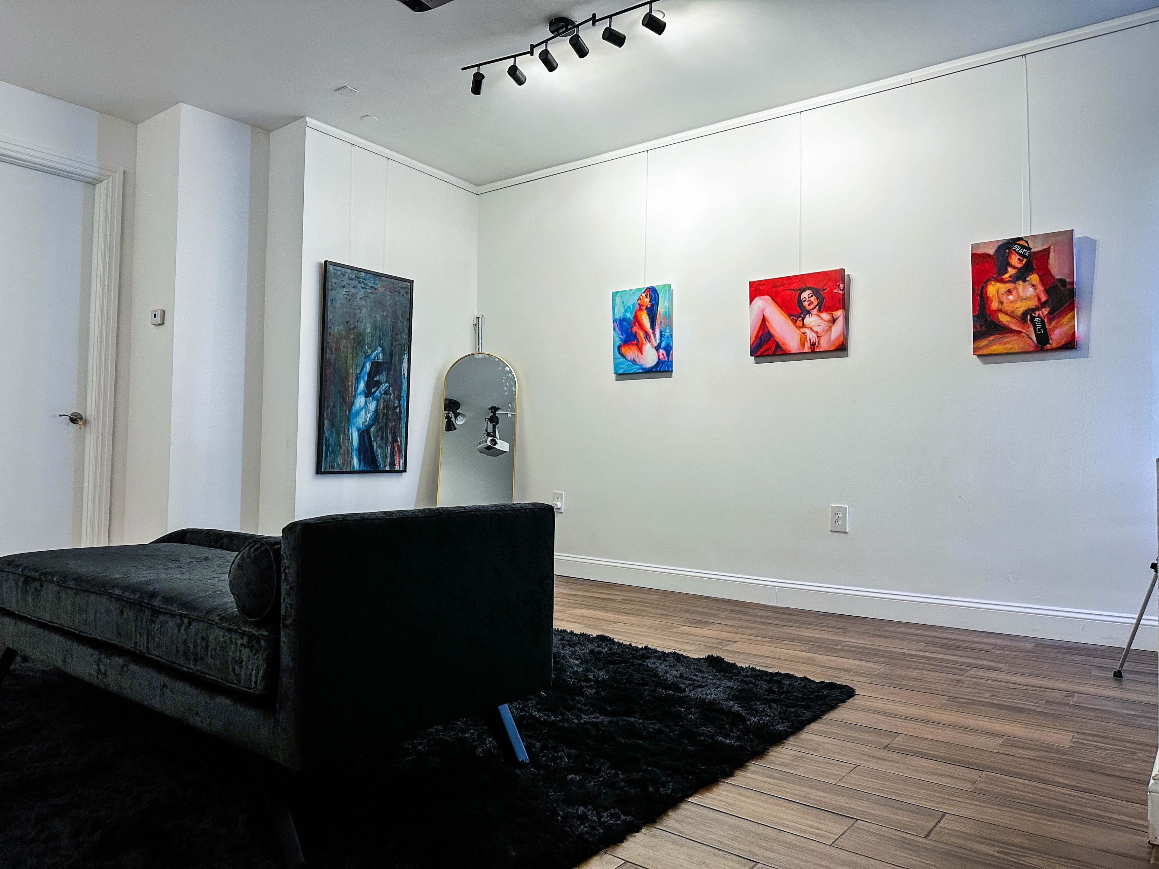 The image shows a modern gallery space featuring four paintings on the wall, a black chaise lounge, and a full-length mirror.