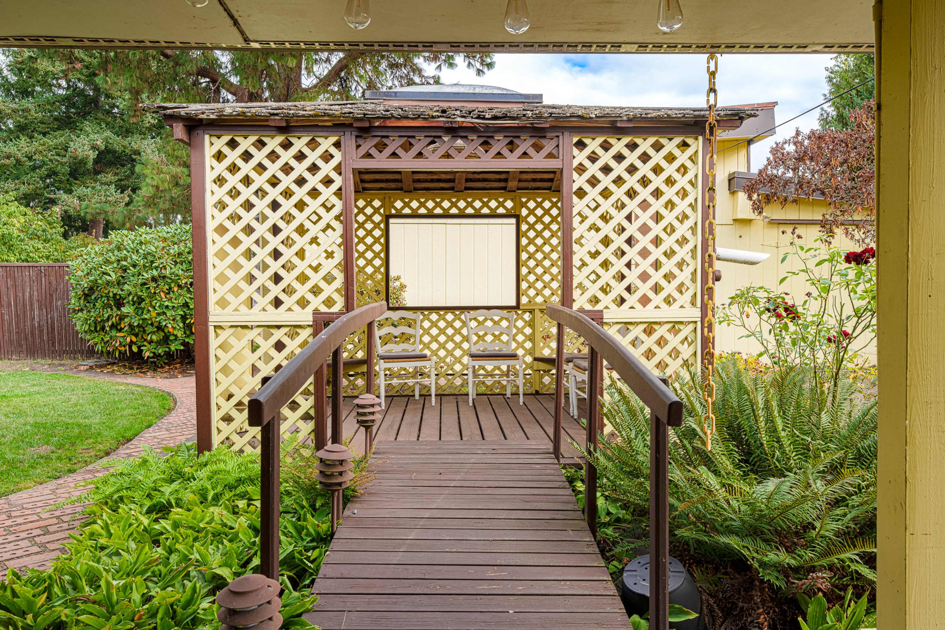 A wooden walkway leads to a lattice-patterned structure surrounded by greenery and a brick path.