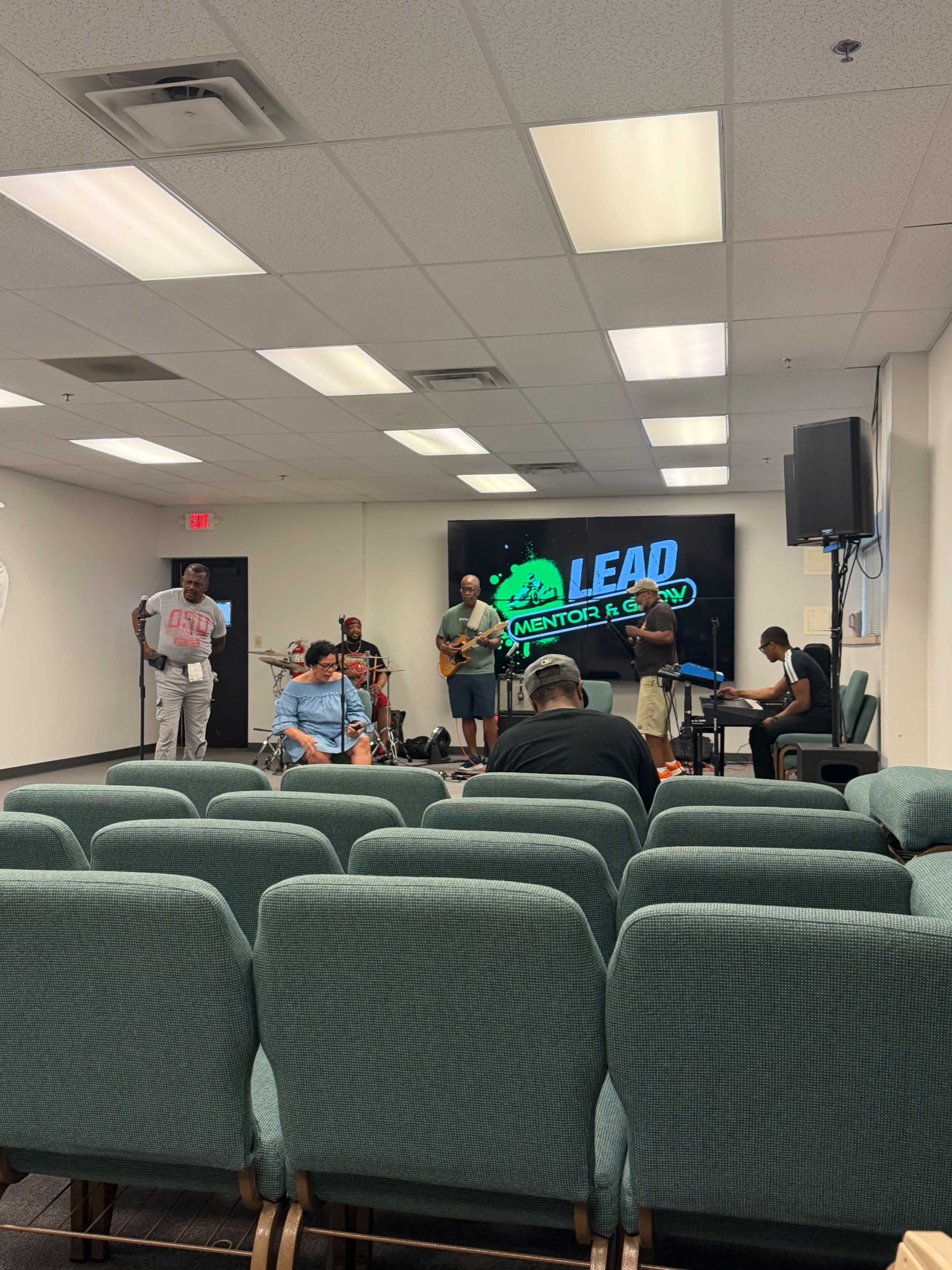 A group of musicians is practicing in a room with chairs arranged in rows and a large screen displaying the word "LEAD."