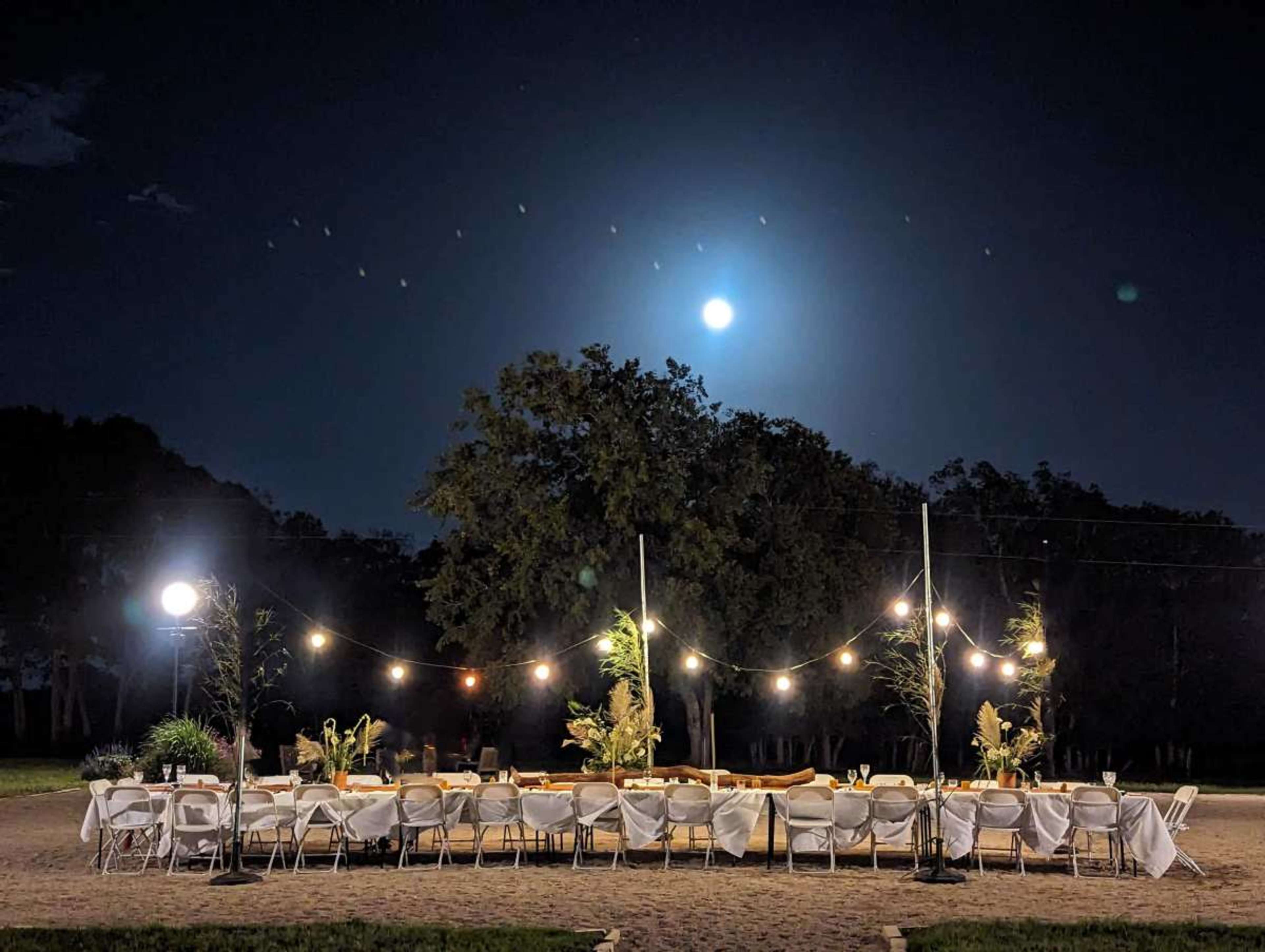 A long table set with chairs is illuminated by string lights under a full moon at night.