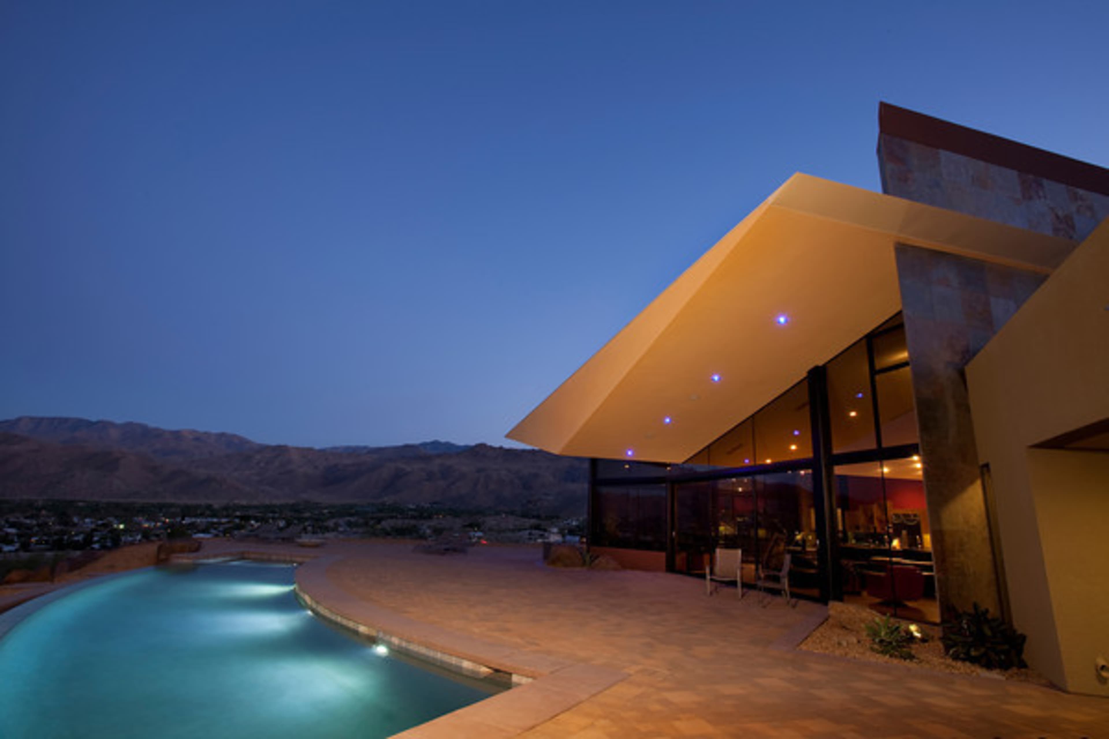 A modern-style home with a large pool is situated against a backdrop of mountains at dusk.