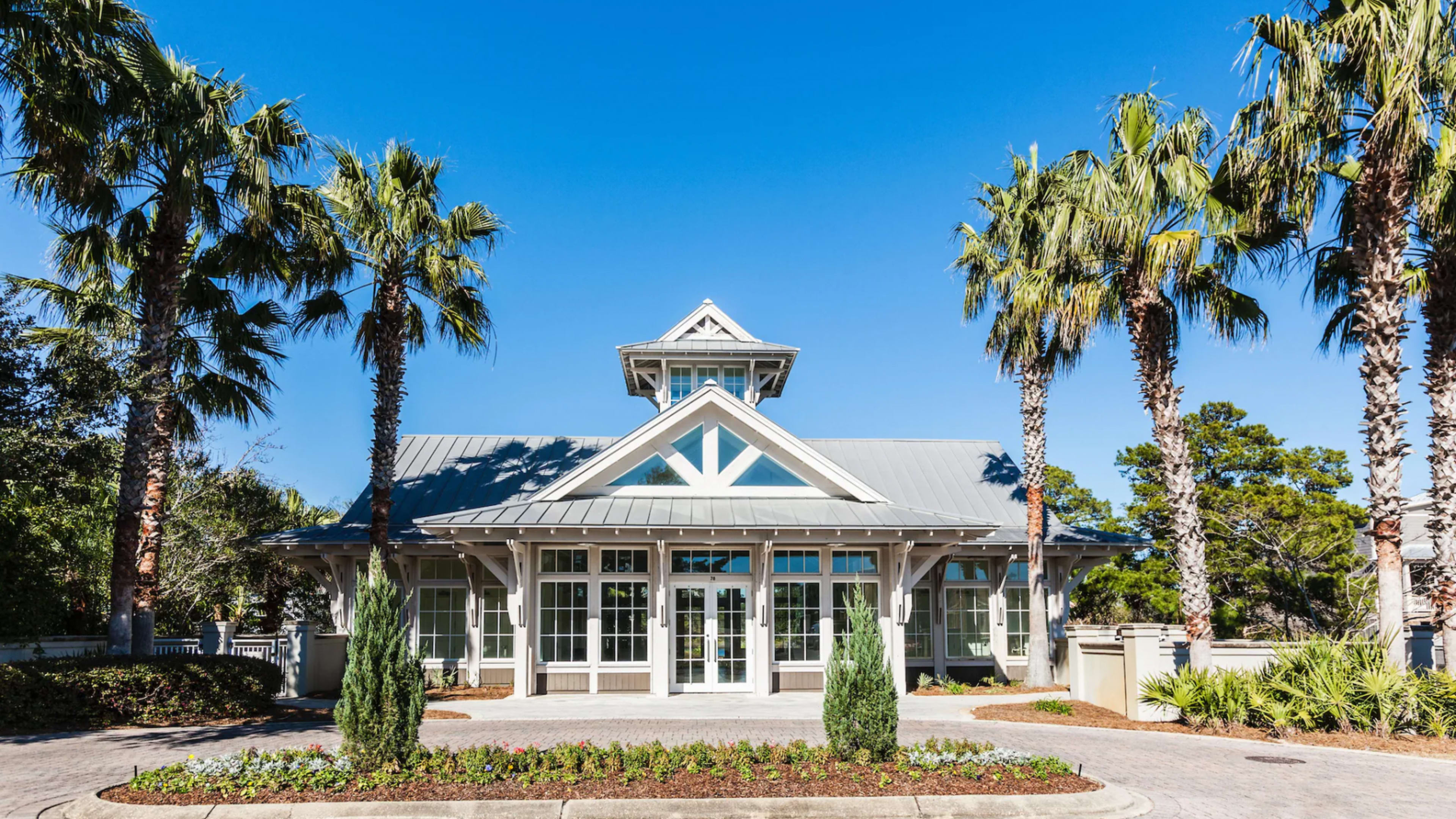 A modern building with a gray metal roof, large windows, and palm trees flanks the entrance.