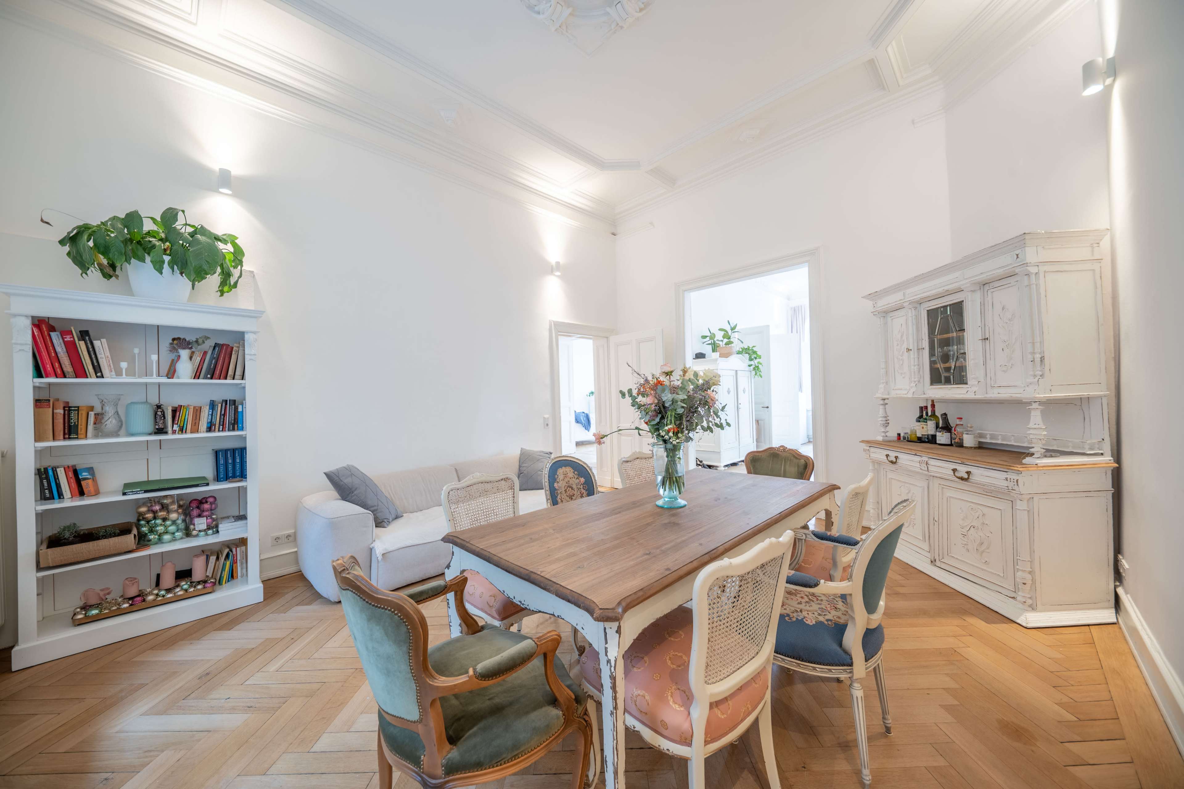 The image shows a bright dining area featuring a wooden table surrounded by several chairs, a bookshelf filled with books, and a decorative sideboard.