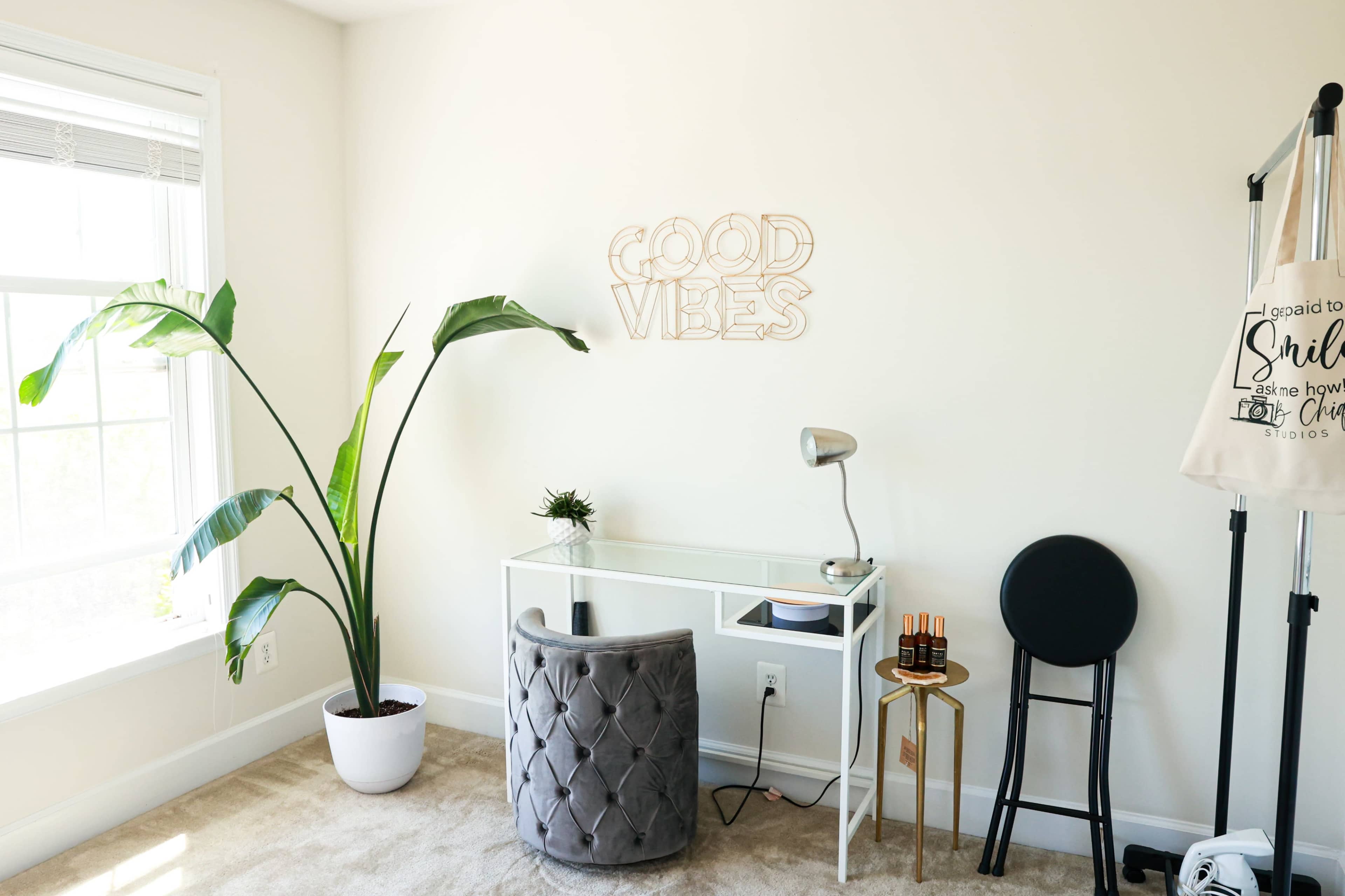 The image shows a minimalist room with a small desk, a chair, a tall indoor plant, and a wall decoration that says "GOOD VIBES."
