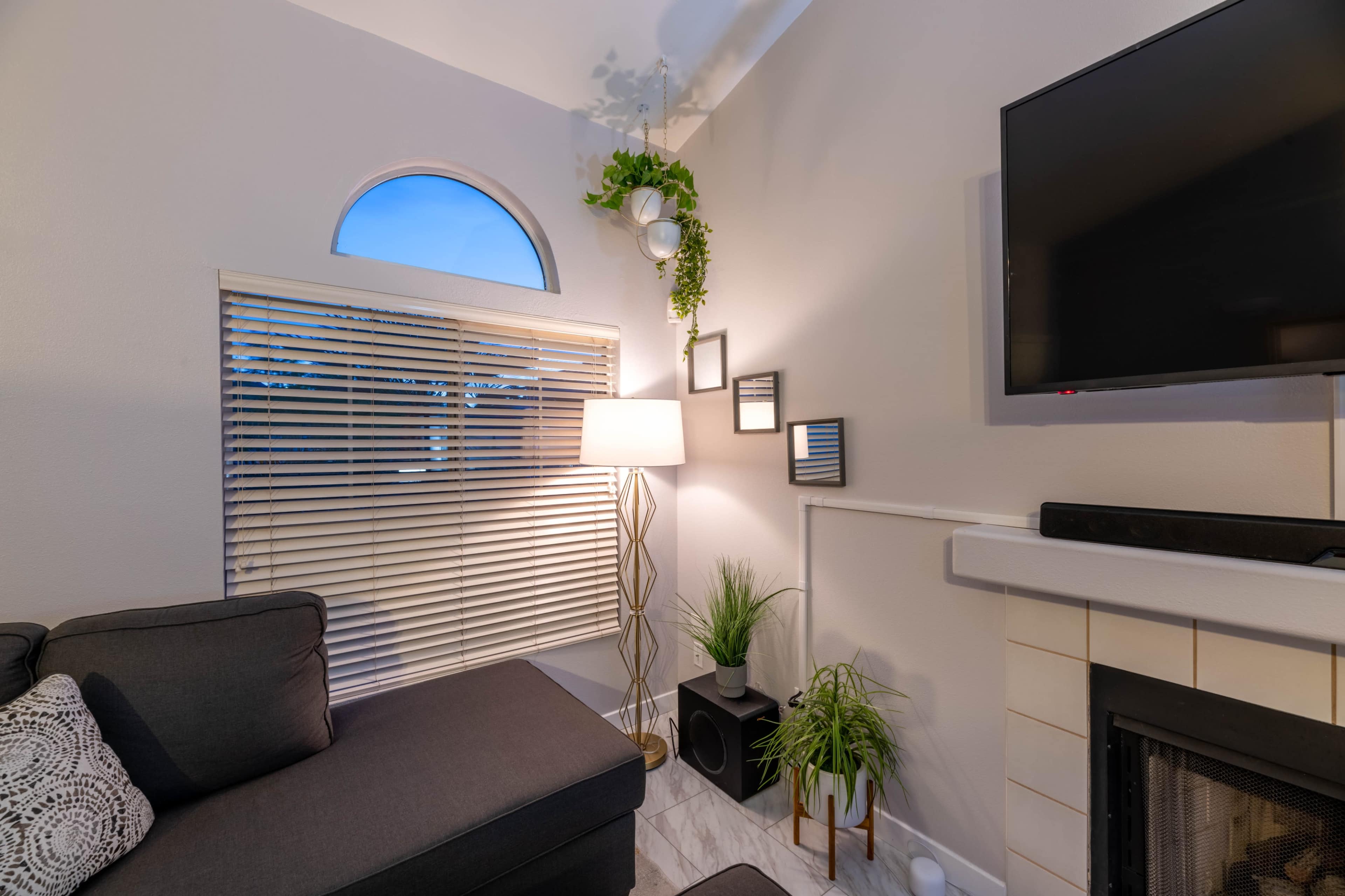A modern living room features a sectional sofa, a floor lamp, a TV mounted on the wall, and decorative plants beside a window with blinds.