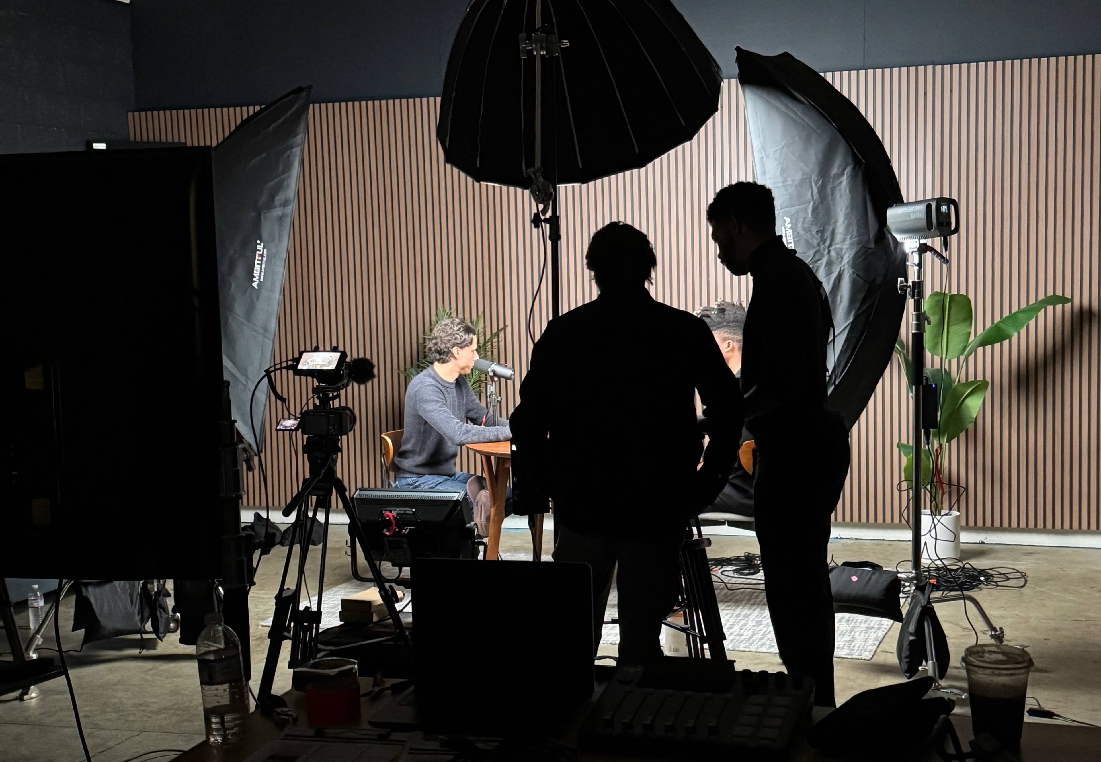 Two individuals are seated at a table in a studio, silhouetted against a backdrop of wooden panels, surrounded by filming equipment and lights.