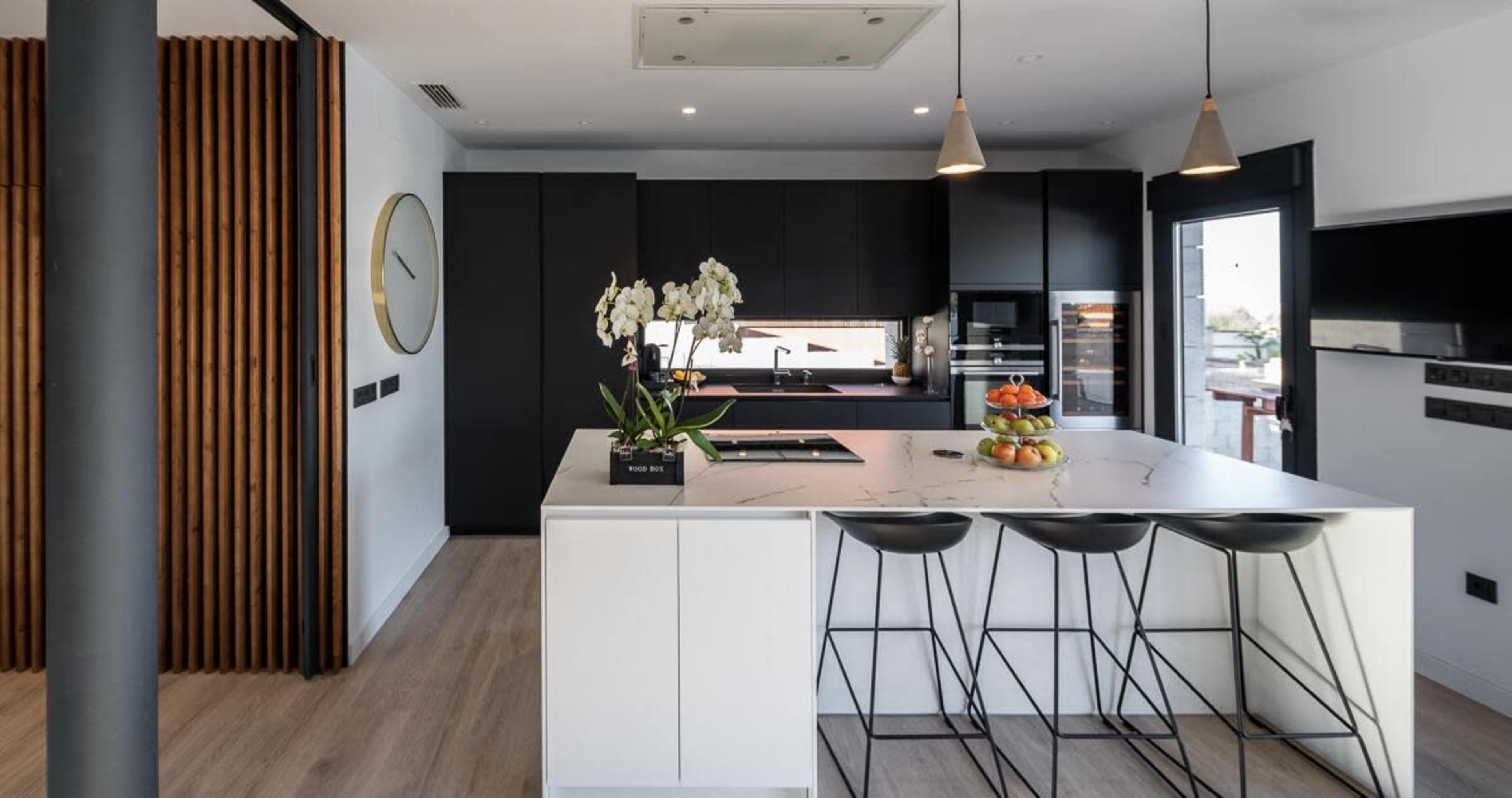 The image shows a modern kitchen featuring a central island with bar stools, dark cabinetry, a wall clock, and large windows.