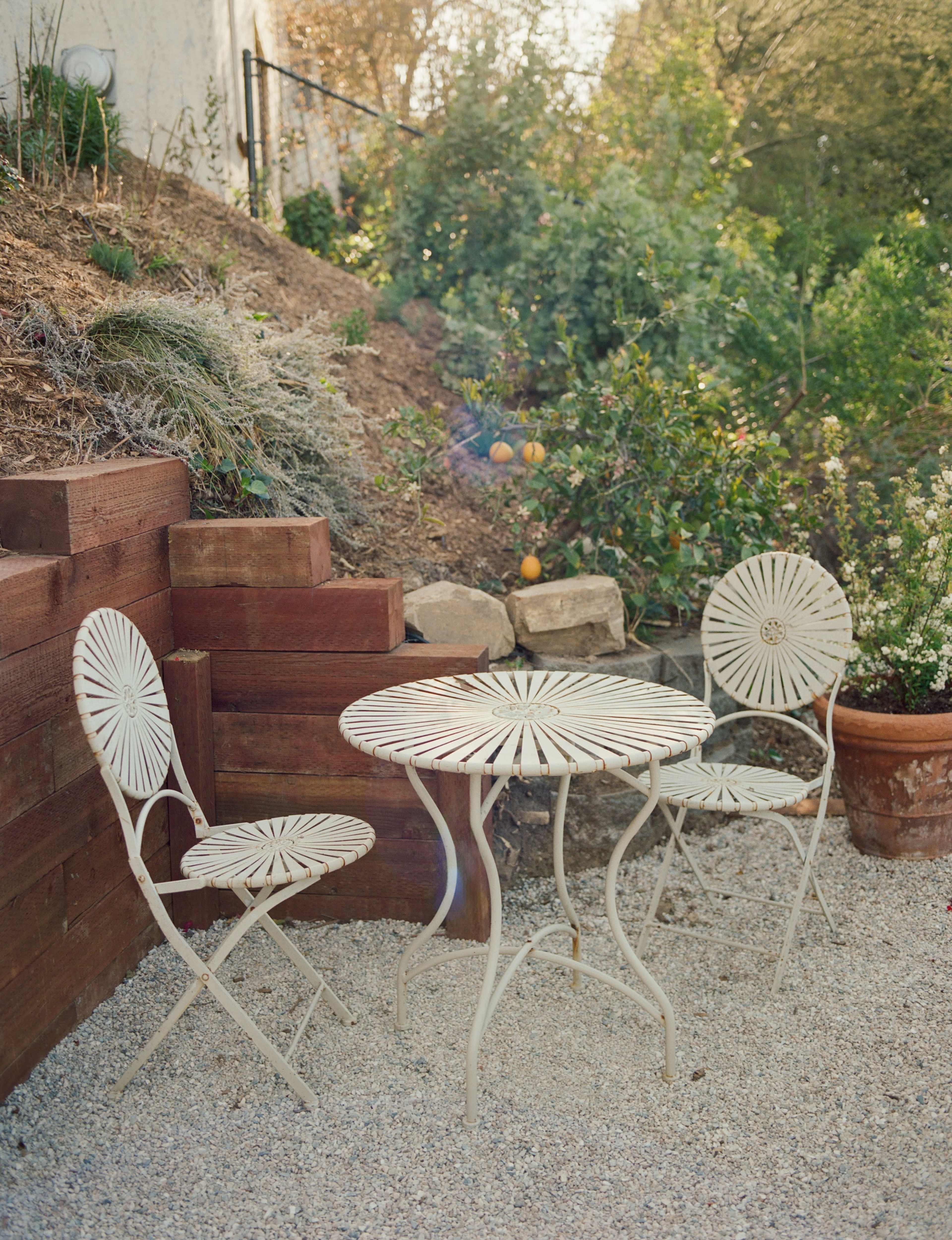 A round table with two matching chairs is set on a gravel patio, surrounded by wooden stairs and greenery.