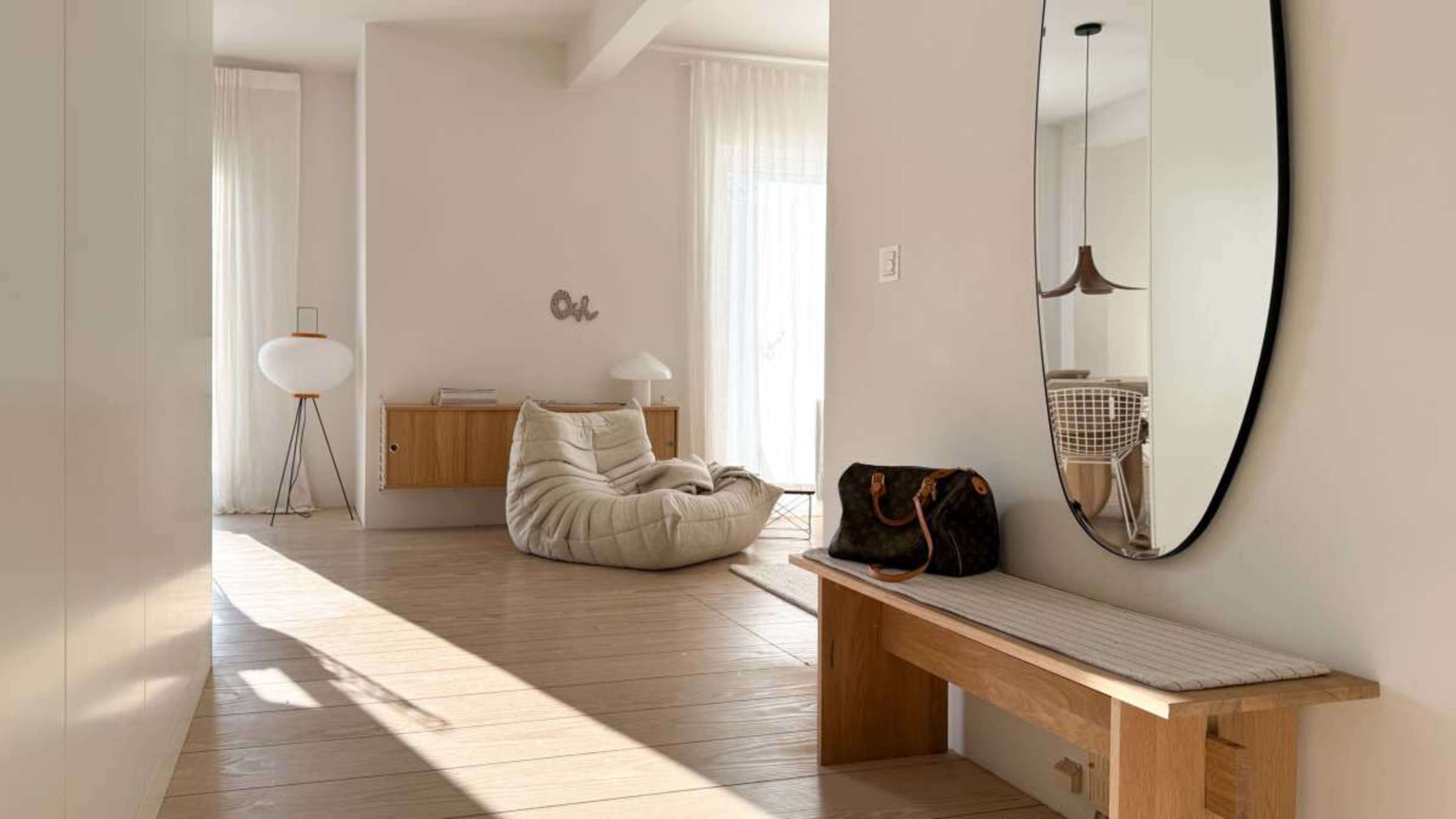 The image shows a spacious, light-filled interior with a wooden bench, a large mirror, and a cozy bean bag chair against a pale wall.