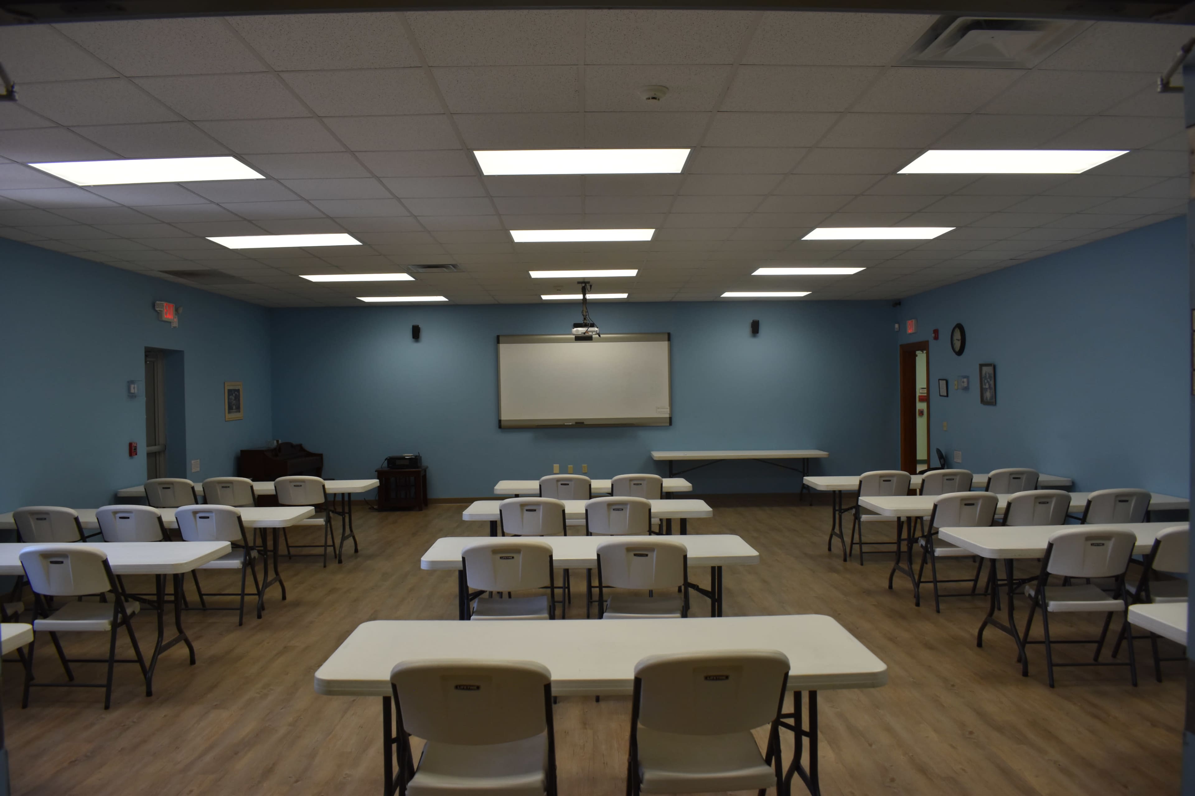 A classroom is arranged with several rows of tables and chairs, featuring blue walls and a whiteboard at the front.