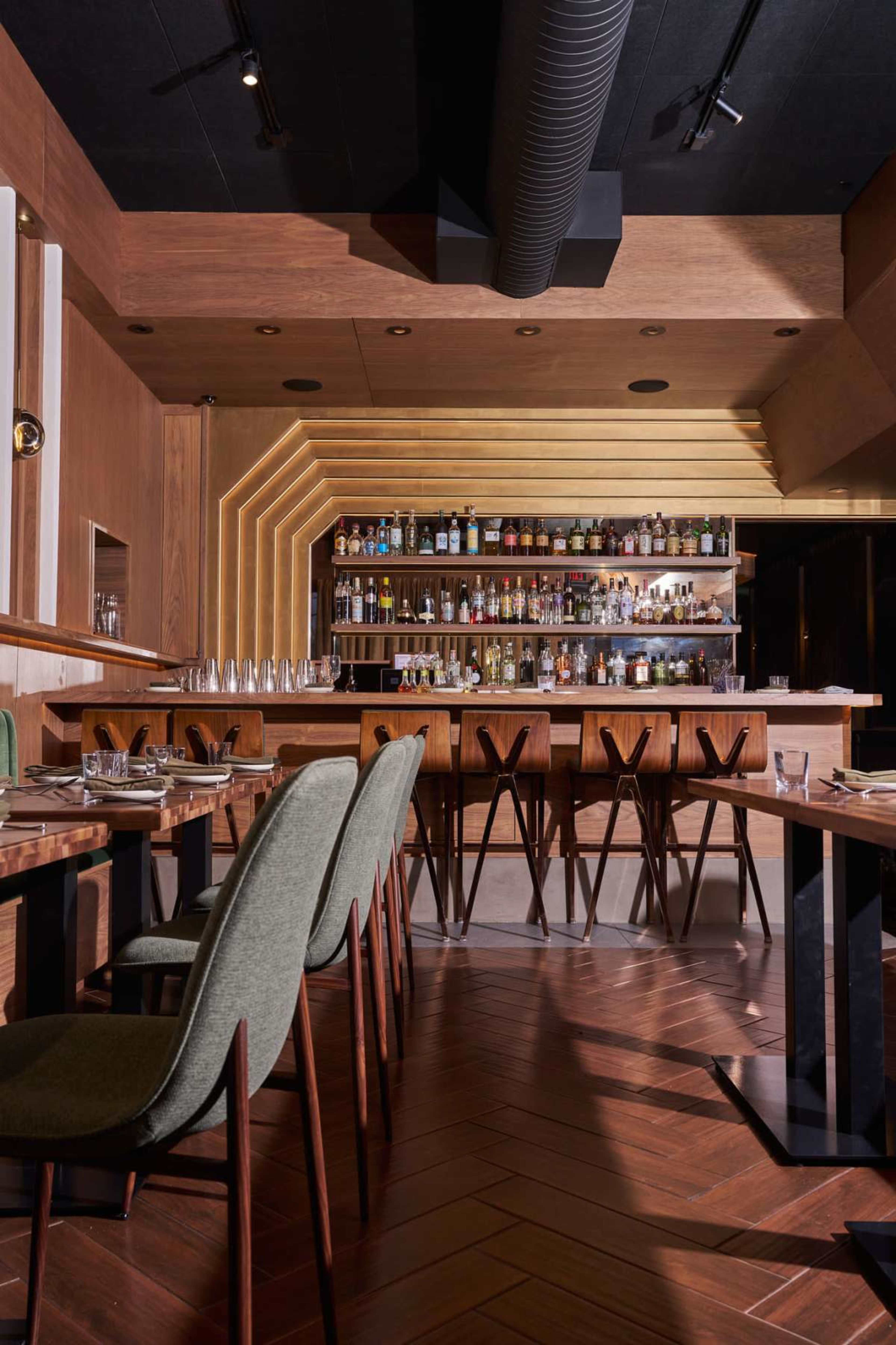 The image shows an interior view of a modern restaurant featuring wooden decor, a bar stocked with various bottles, and tables with green upholstered chairs.