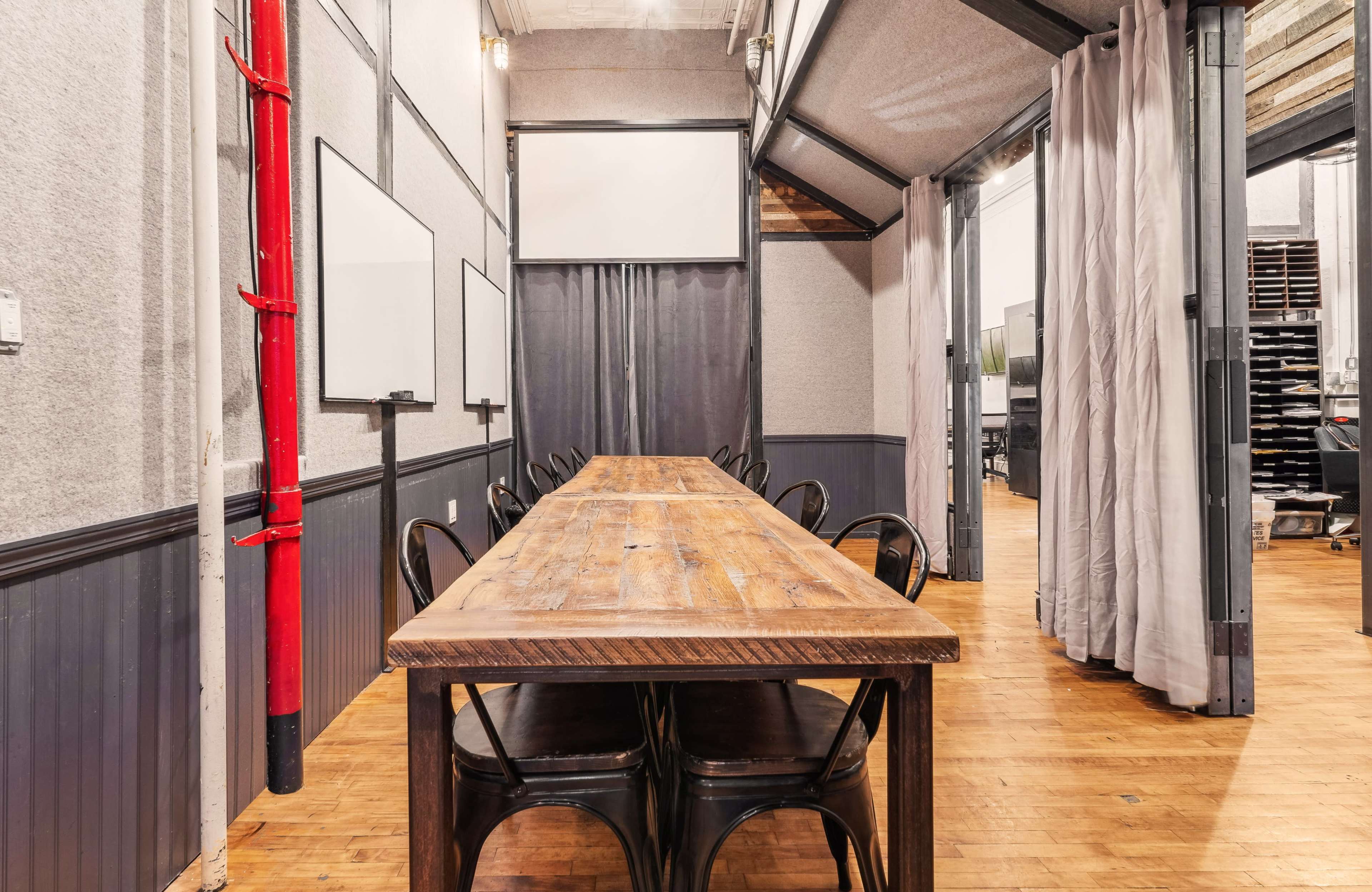 A long wooden table with metal chairs is positioned in a meeting room featuring gray walls and sliding curtains.