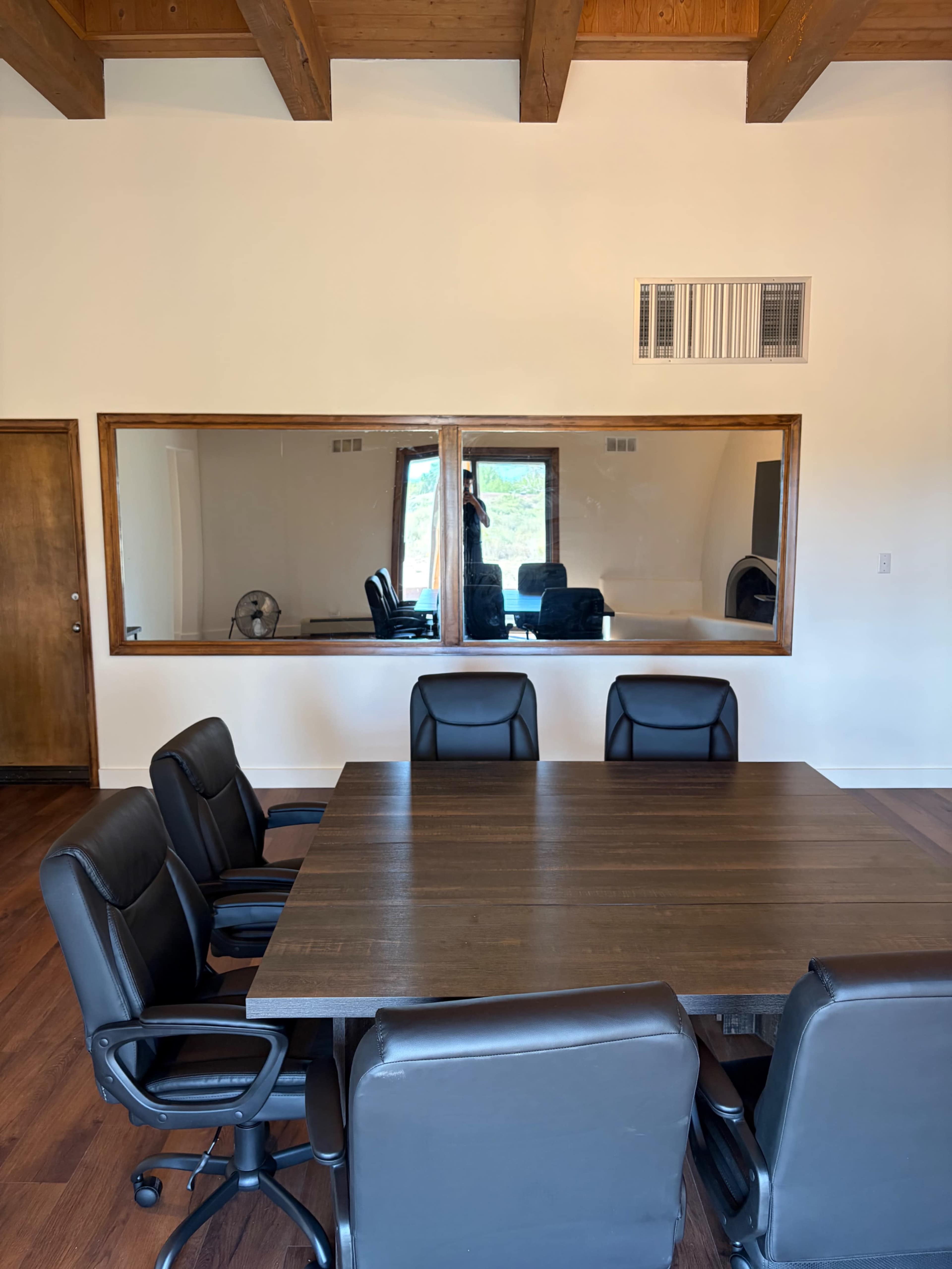 A conference room features a large wooden table surrounded by black leather chairs, with a large mirrored window reflecting a separate space.