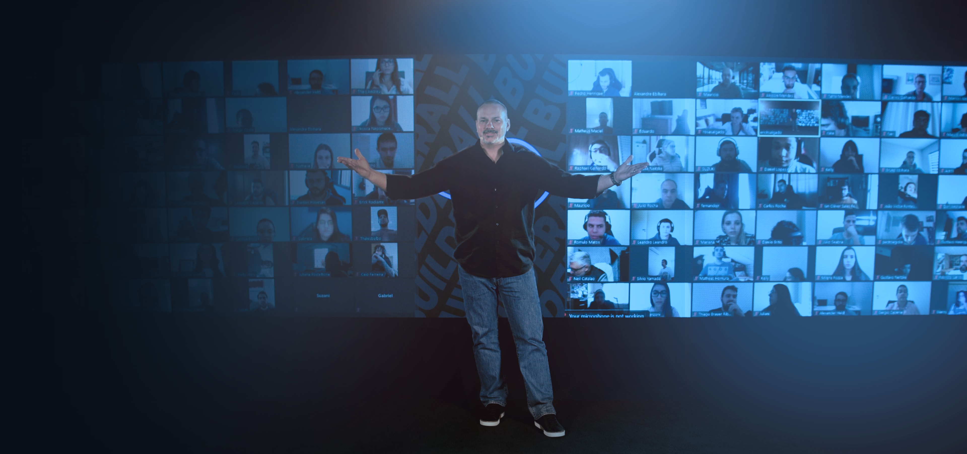 A man stands in front of a large screen displaying multiple video call participants.