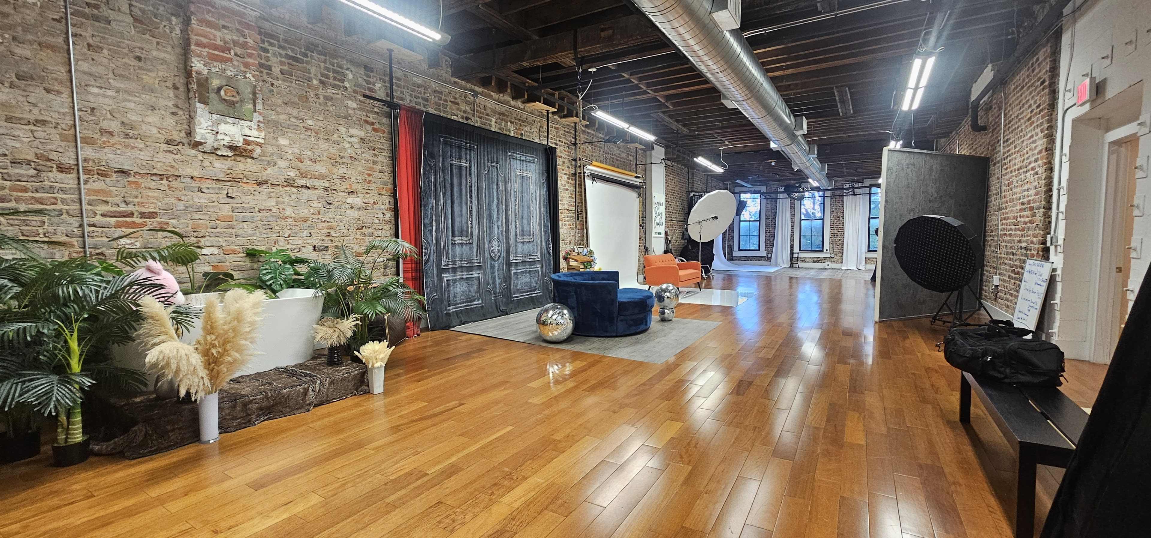 The image shows a spacious studio with brick walls, wooden flooring, and various decorative elements including plants, a colorful sofa, and photography props.