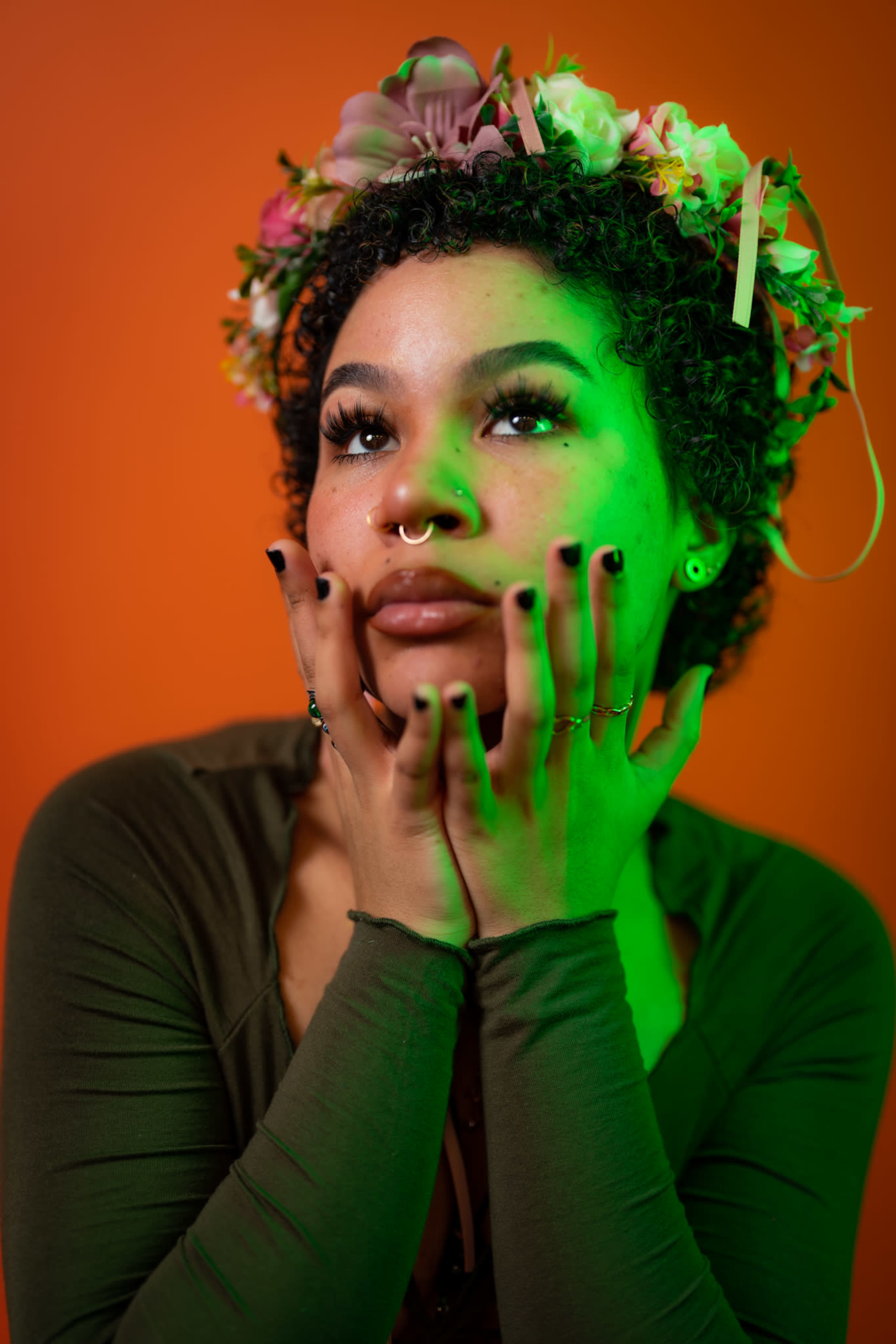 A person with curly hair and a flower crown poses against an orange background, resting their hands on their face under a green light.