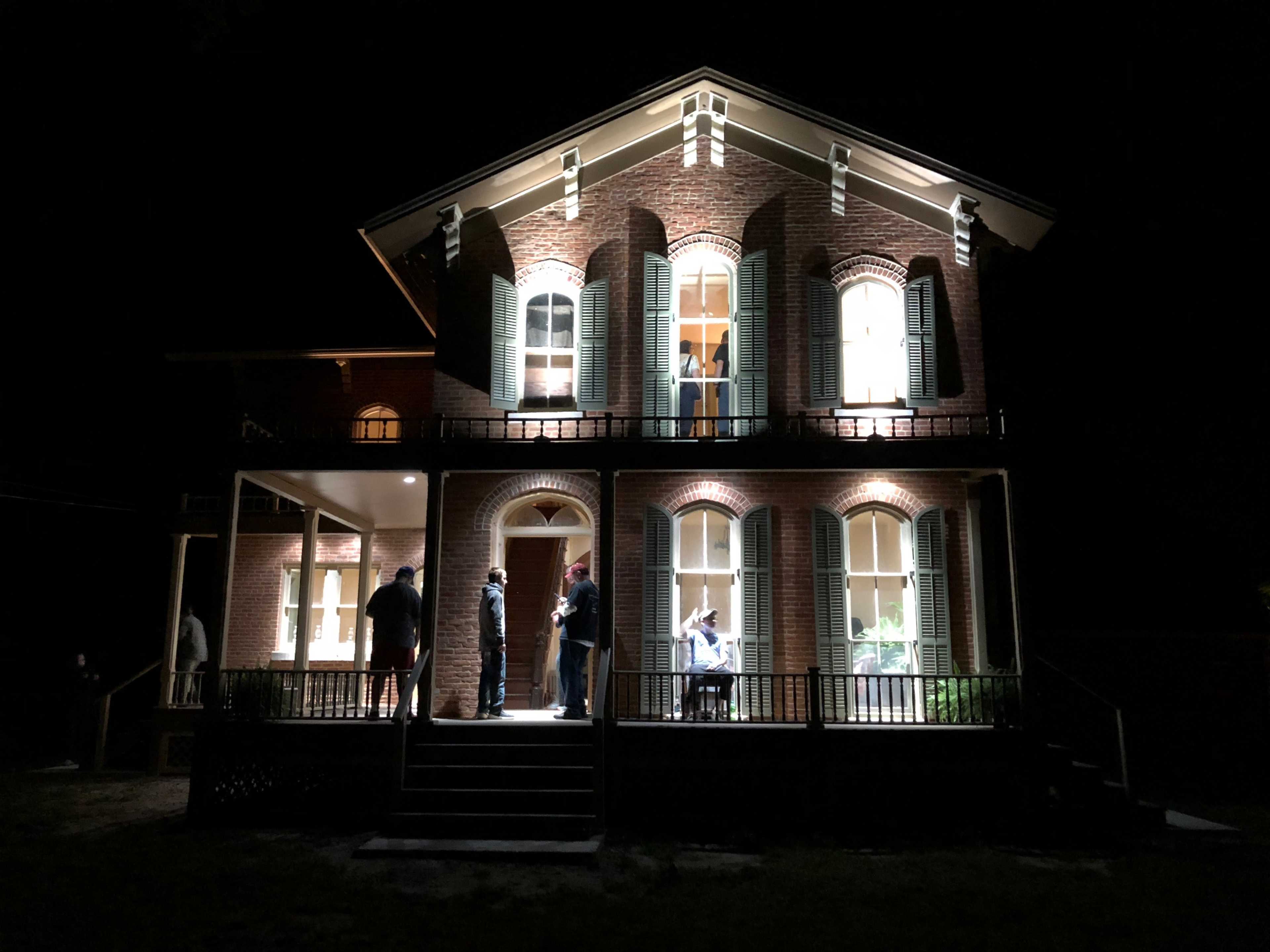 A well-lit, two-story brick house is illuminated at night, with several people gathered on the porch and steps.