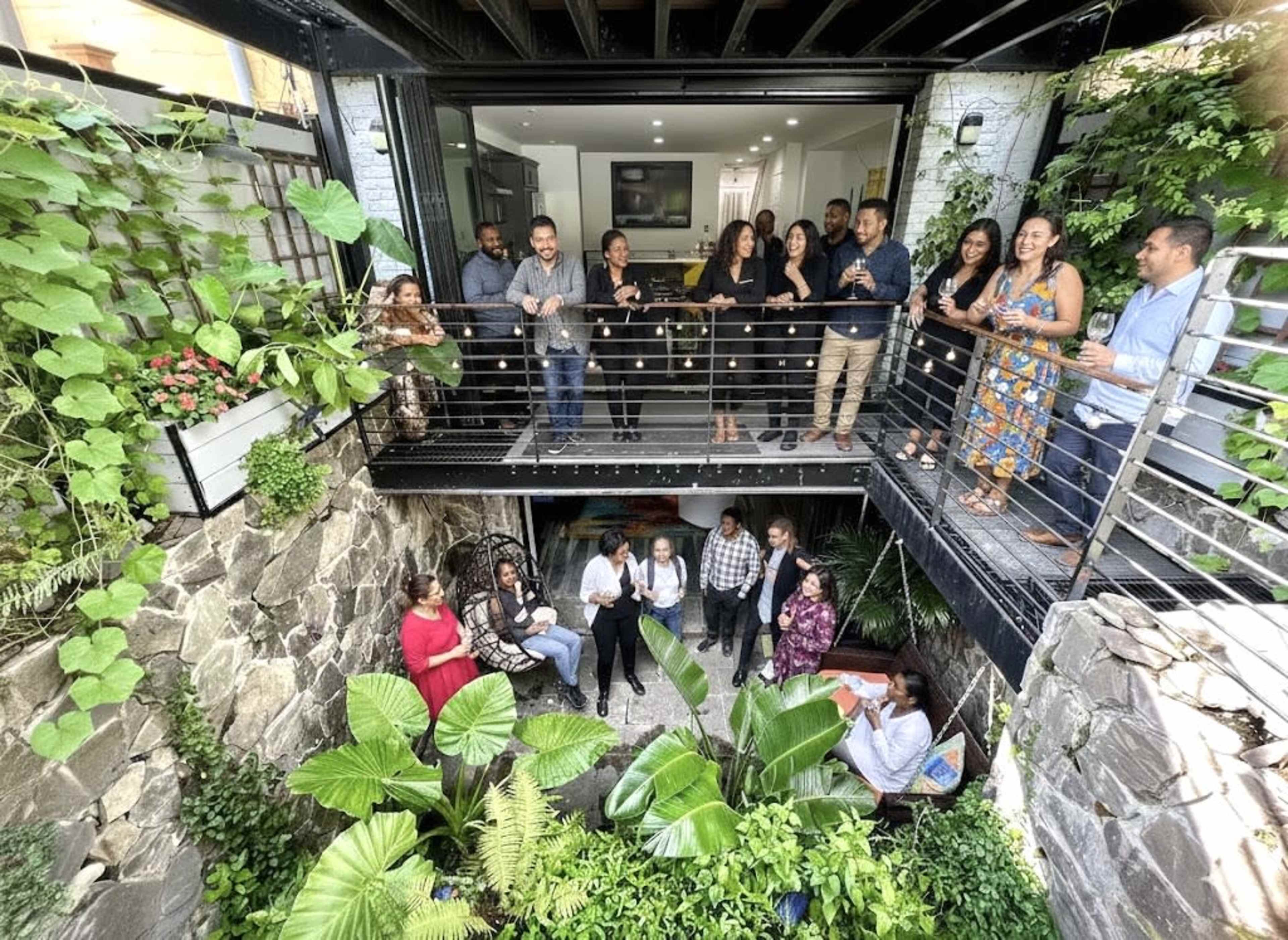 A group of people stands on a balcony overlooking a green courtyard filled with plants and a few seated individuals.